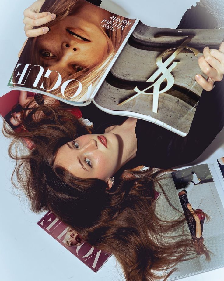 Woman lying down holding Vogue magazine with YSL advertisement, surrounded by fashion magazines on white surface.