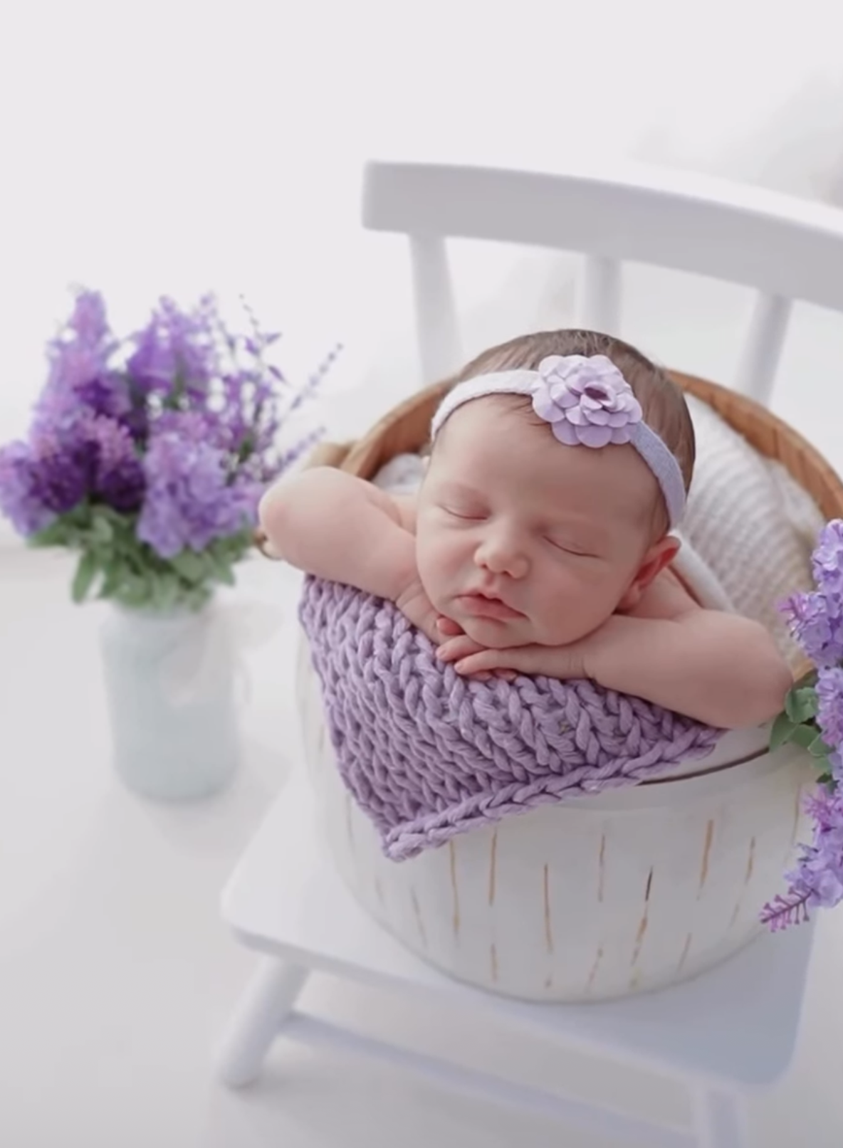 Sleeping newborn baby girl in white basket with purple knit blanket and floral headband, surrounded by lavender flowers.