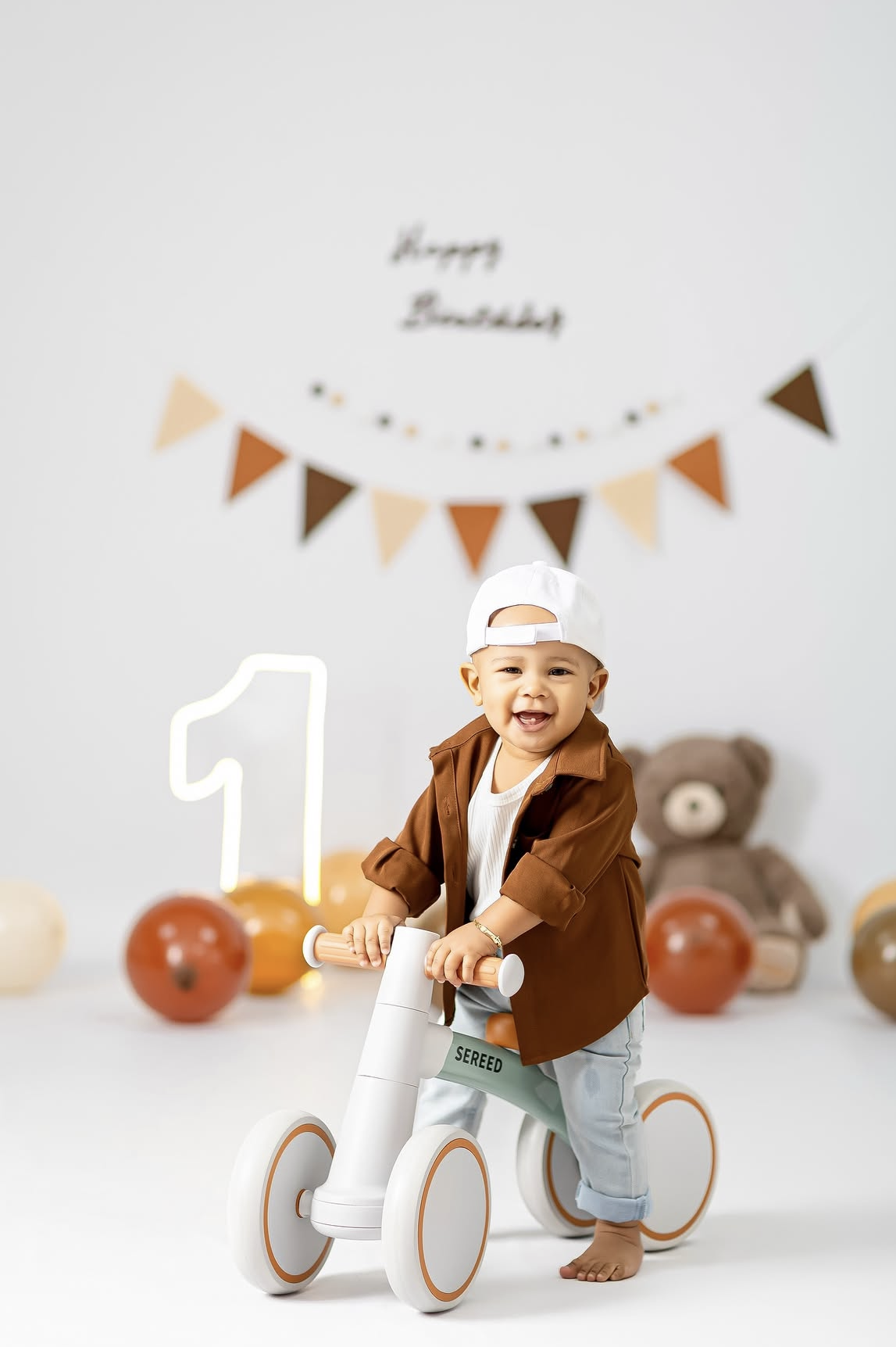 Smiling toddler on white balance bike celebrating 1st birthday with decorations, balloons, and teddy bear in background.