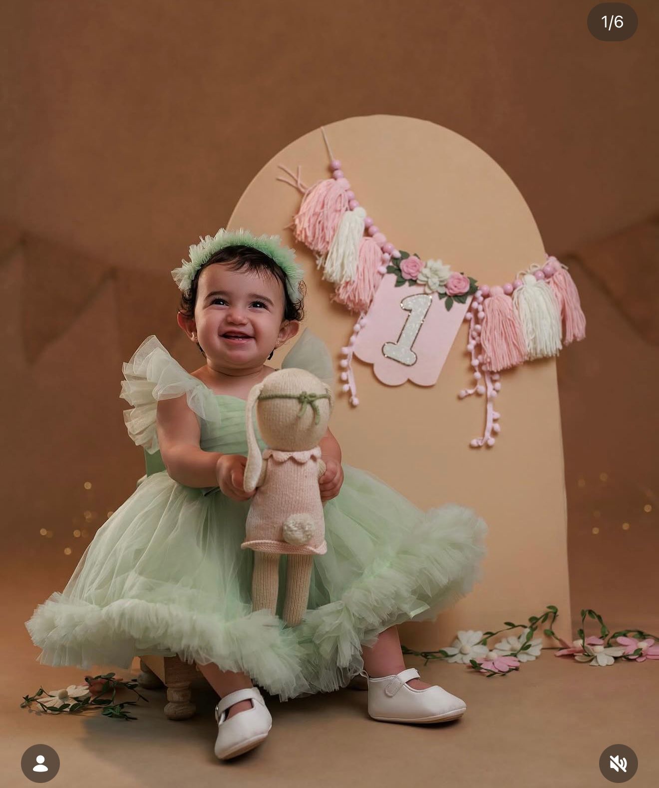 Baby girl in mint green tutu dress holds doll, smiling near a number 1 birthday banner decoration.
