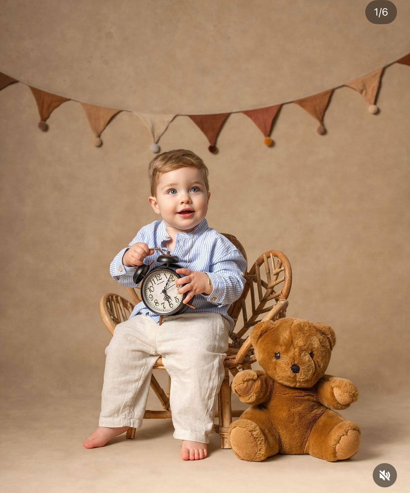 Toddler boy in blue shirt holds alarm clock on rocking chair beside teddy bear, with bunting backdrop.