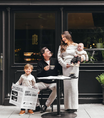 Family enjoying coffee outside a cafe with a baby and toddler holding a newspaper sign.