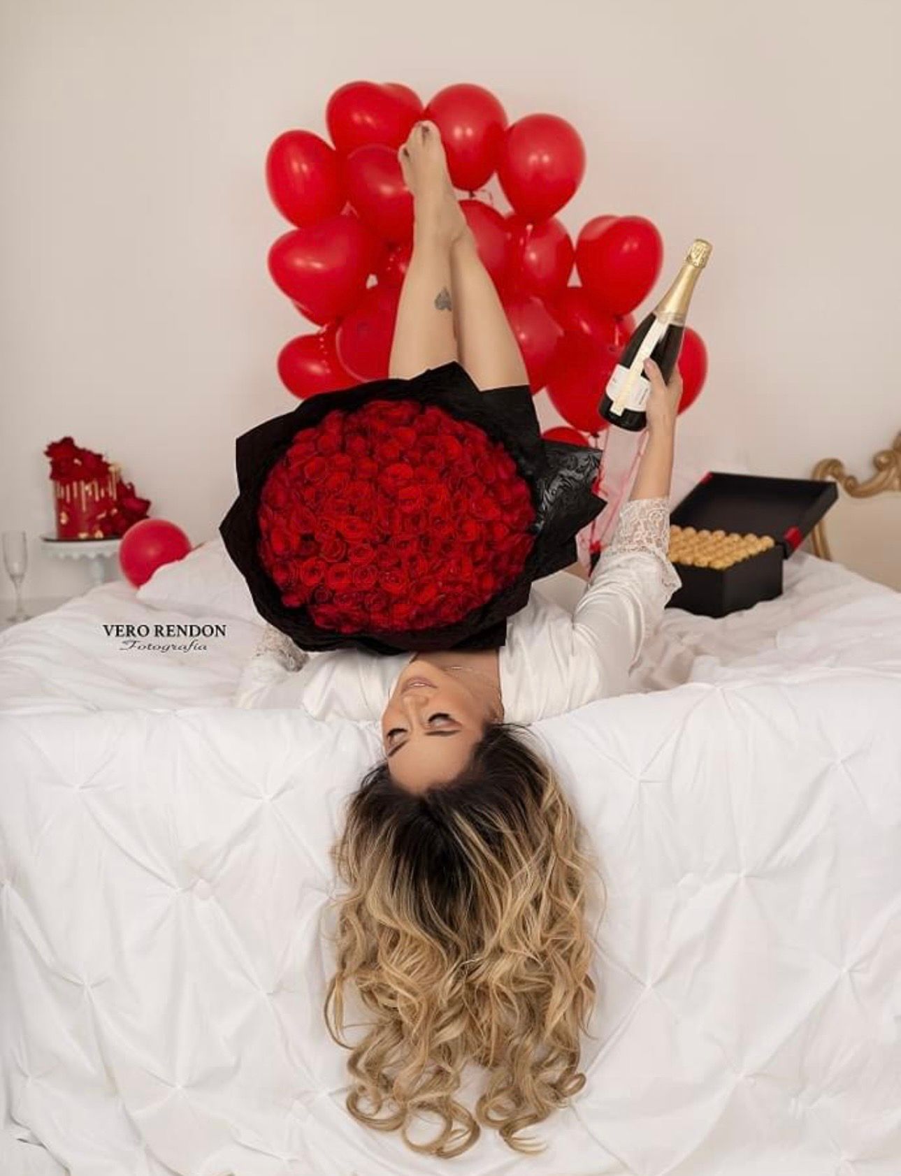 Woman lying on bed holding champagne bottle with red roses bouquet and heart balloons backdrop.