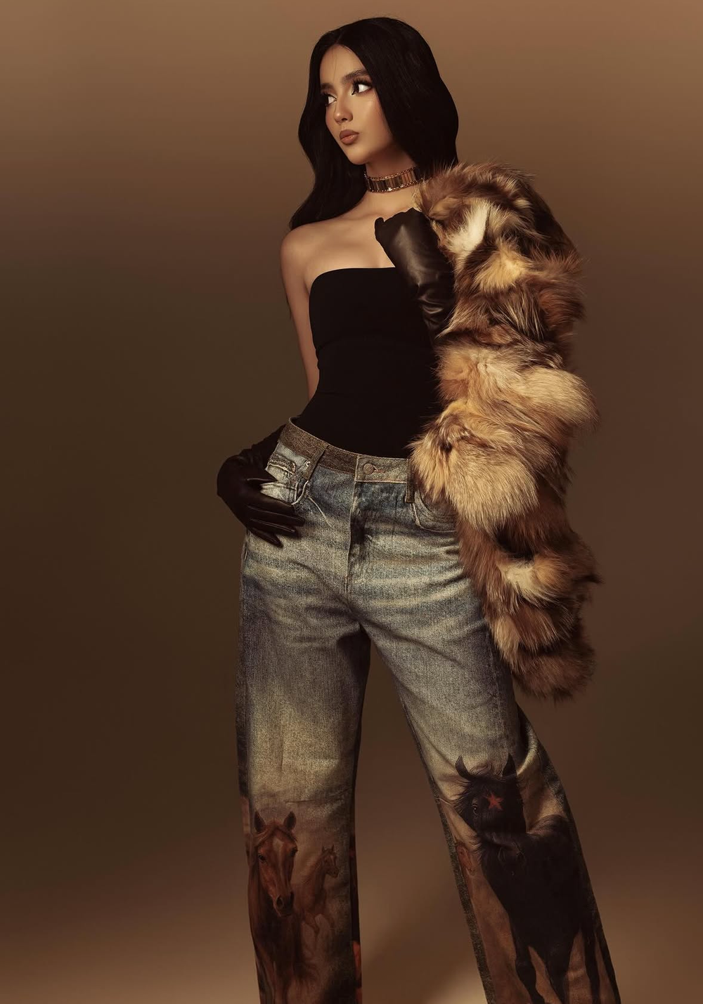 Woman in black strapless top, fur coat draped over shoulder, leather gloves, and printed denim jeans poses confidently.
