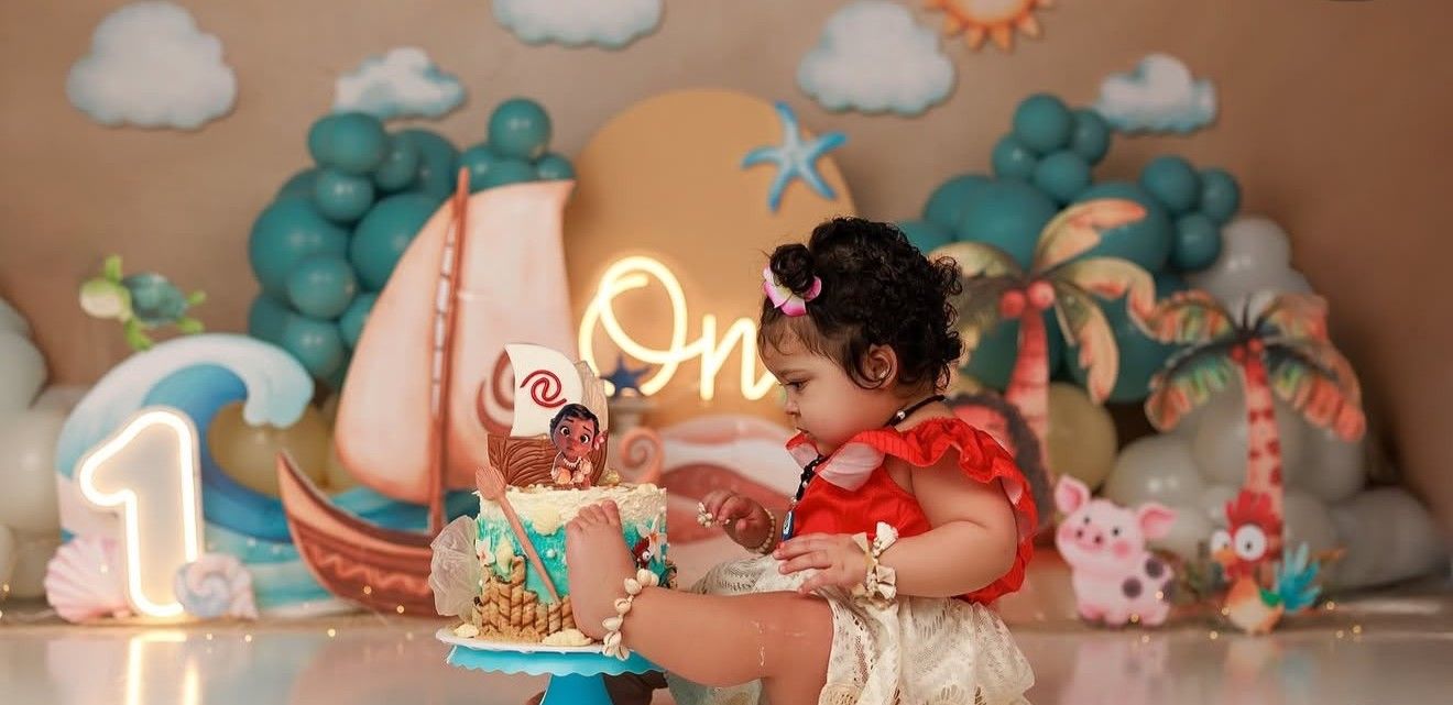 Toddler in red Moana costume smashes first birthday cake at tropical Hawaiian themed party.