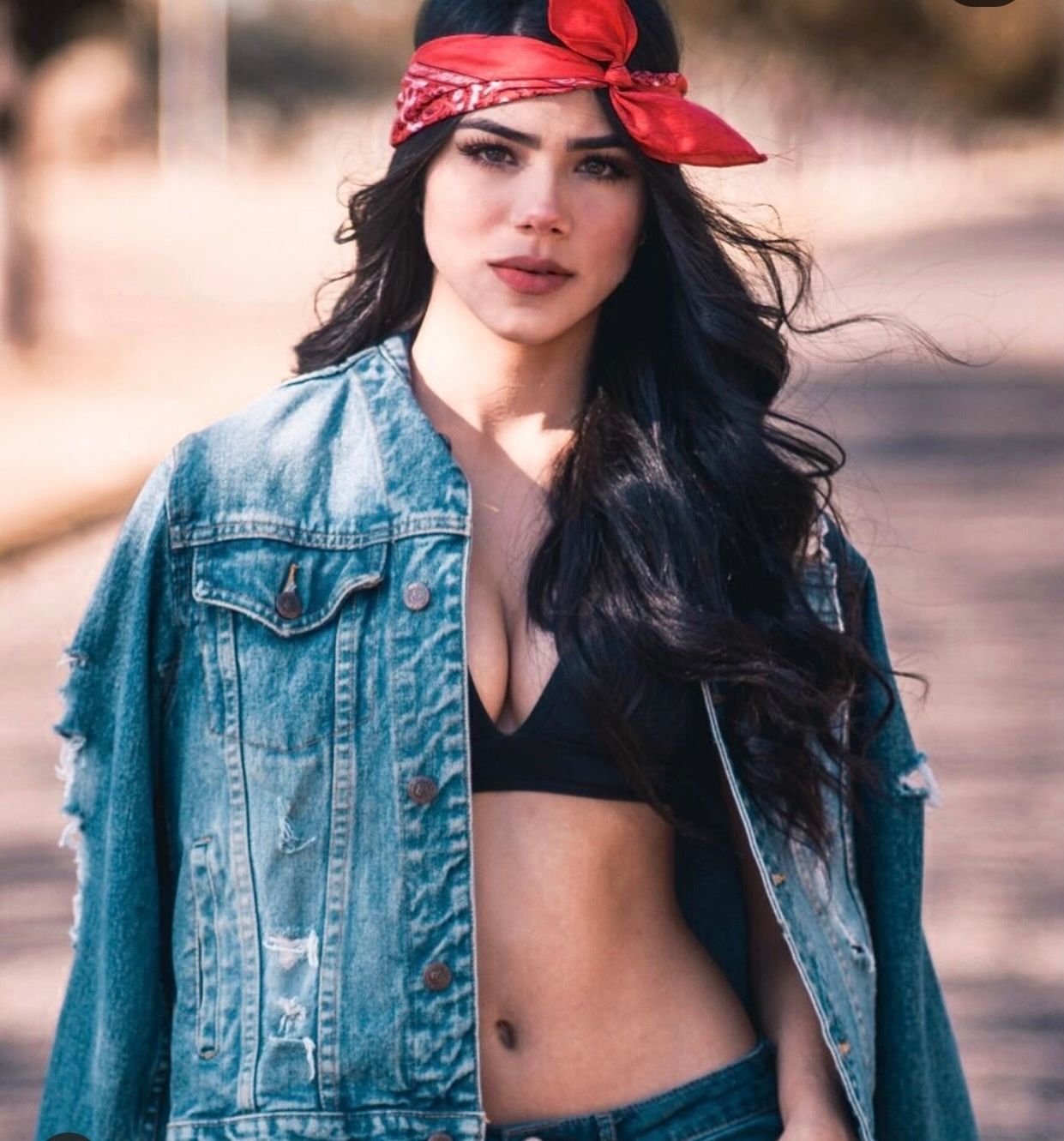 Young woman in denim jacket, black crop top, and red bandana headband poses outdoors with long dark wavy hair.