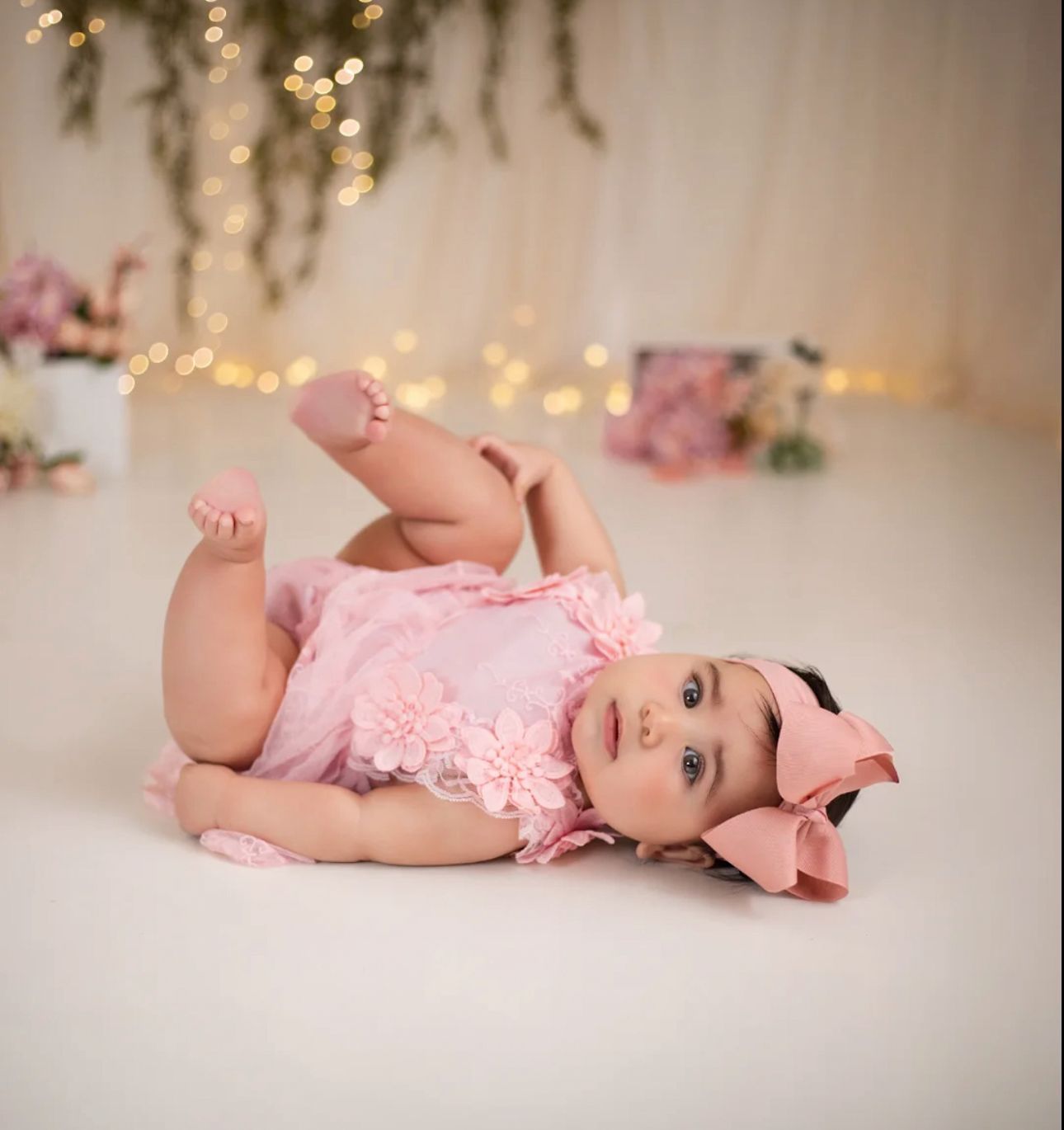 Baby in pink lace romper lying on soft white backdrop with twinkling lights in the background.