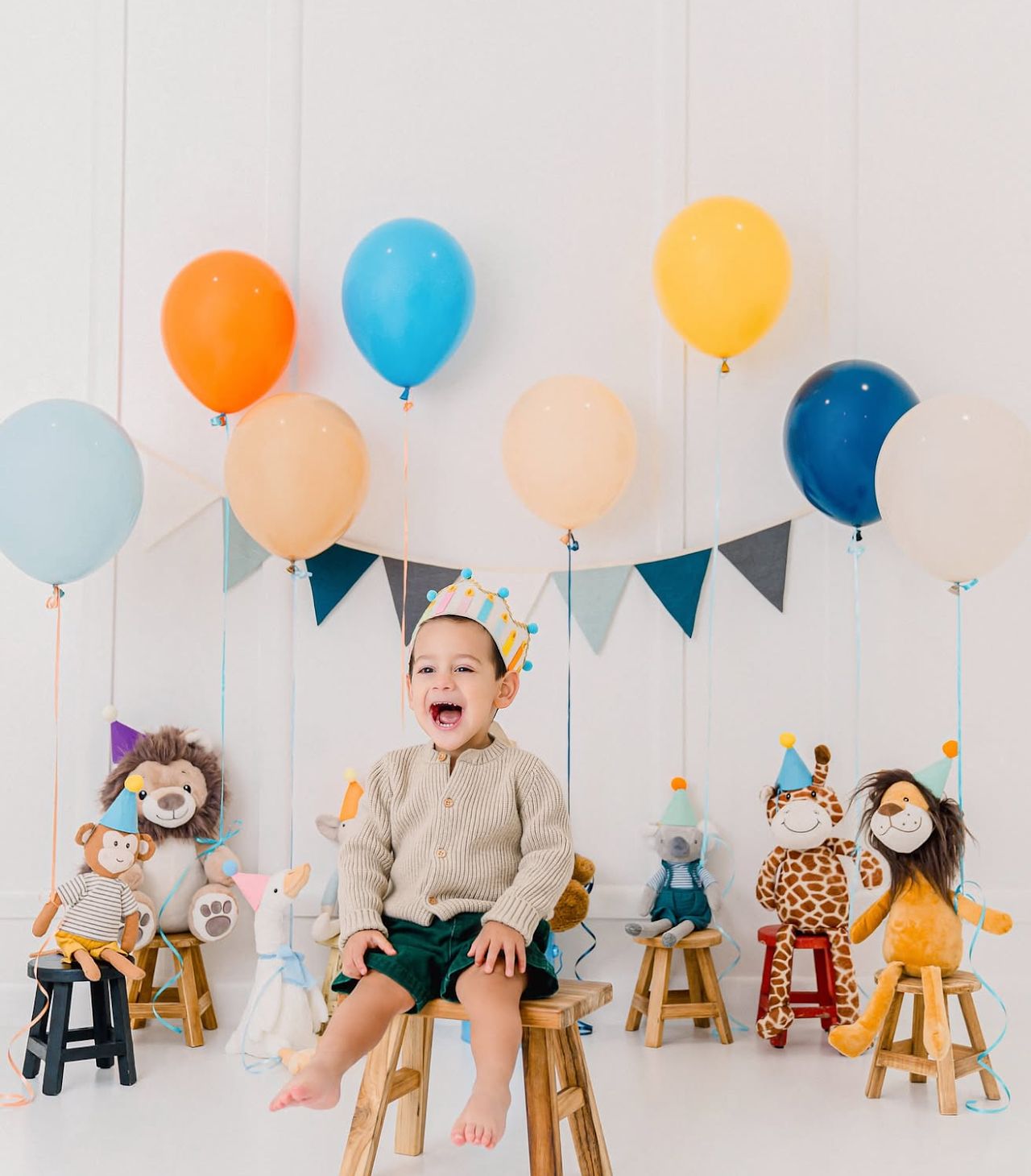 Baby sits on wooden stool with colorful balloons and stuffed animal toys in white studio setting.