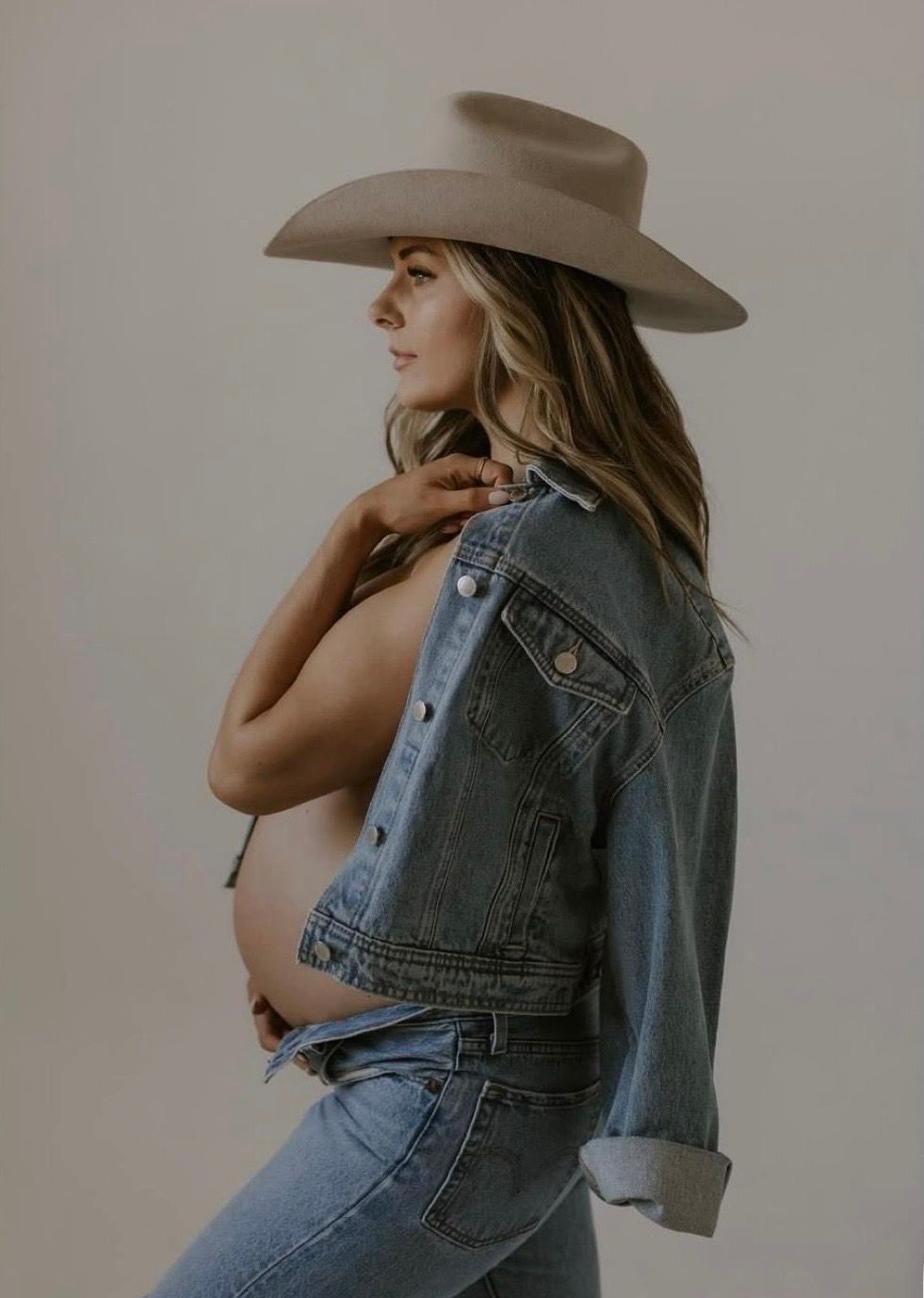 Pregnant woman in denim jacket and wide-brim hat posing for a maternity photo shoot.