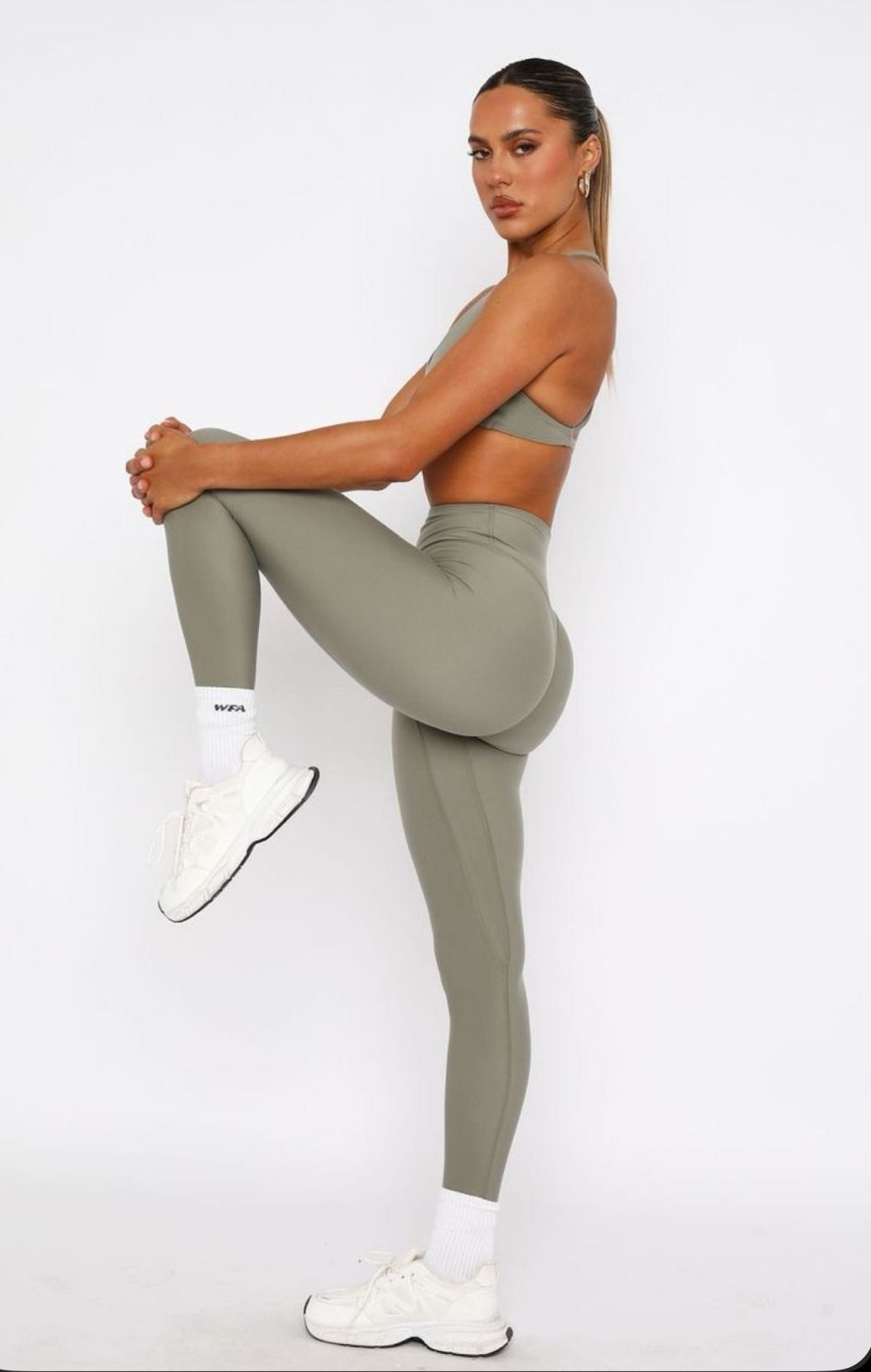 Woman in sage green sports bra and high-waist leggings posing with knee raised, wearing white sneakers and socks.