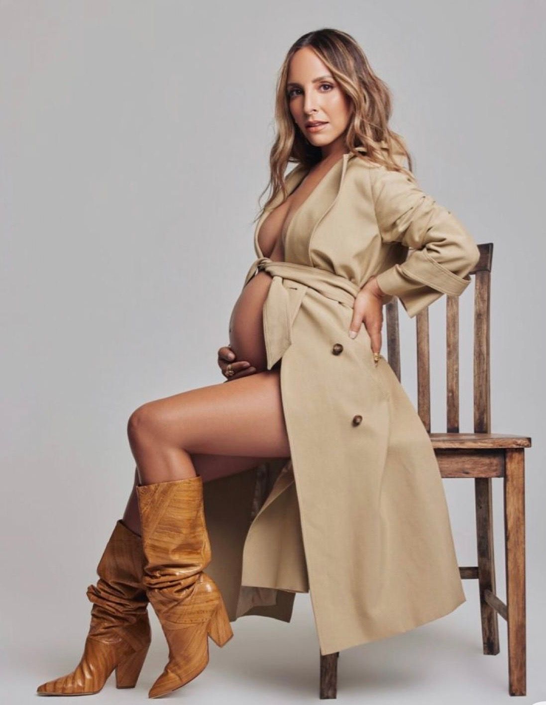 Pregnant woman posing in beige trench coat and brown knee-high boots, seated on wooden chair against gray background.
