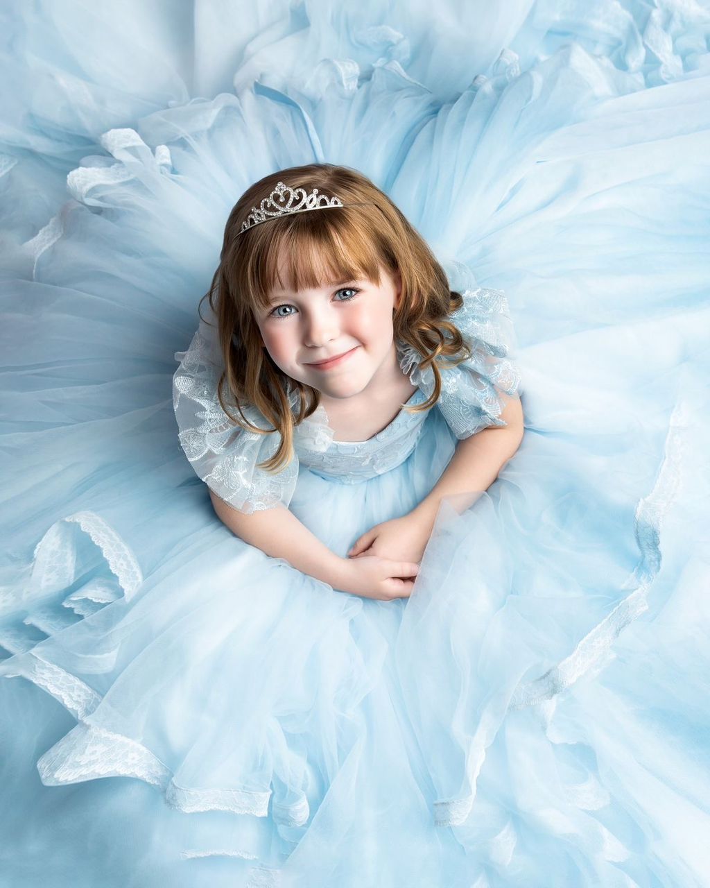 Young girl in a light blue tulle princess gown and silver tiara smiles up at camera in a dreamy portrait.