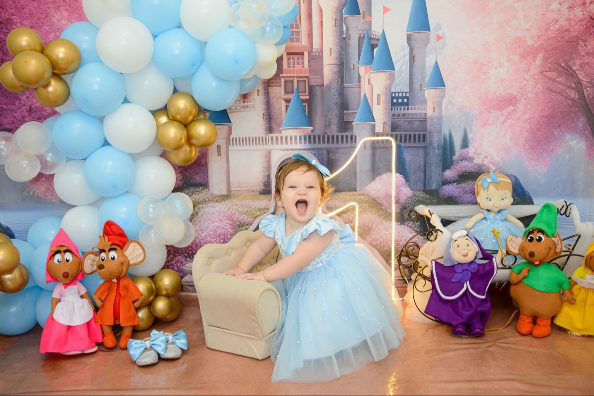 Toddler girl in blue Cinderella dress at princess castle birthday party with character plush toys and balloon decorations.