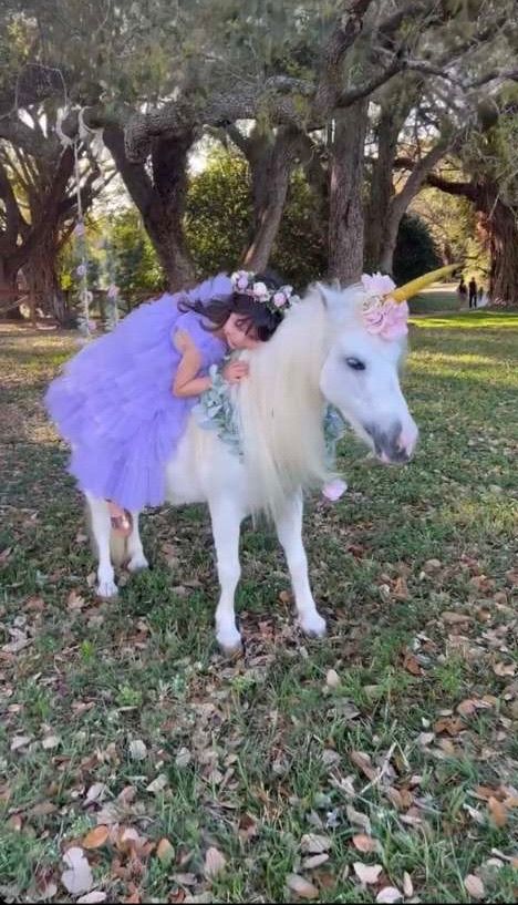 A young girl in a purple tutu dress hugs a white pony dressed as a unicorn with a gold horn in a park.