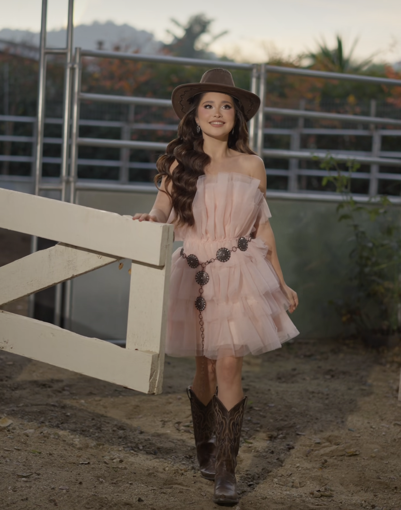 Woman in pink tulle dress, cowboy hat, and boots posing at a ranch fence in golden hour light.