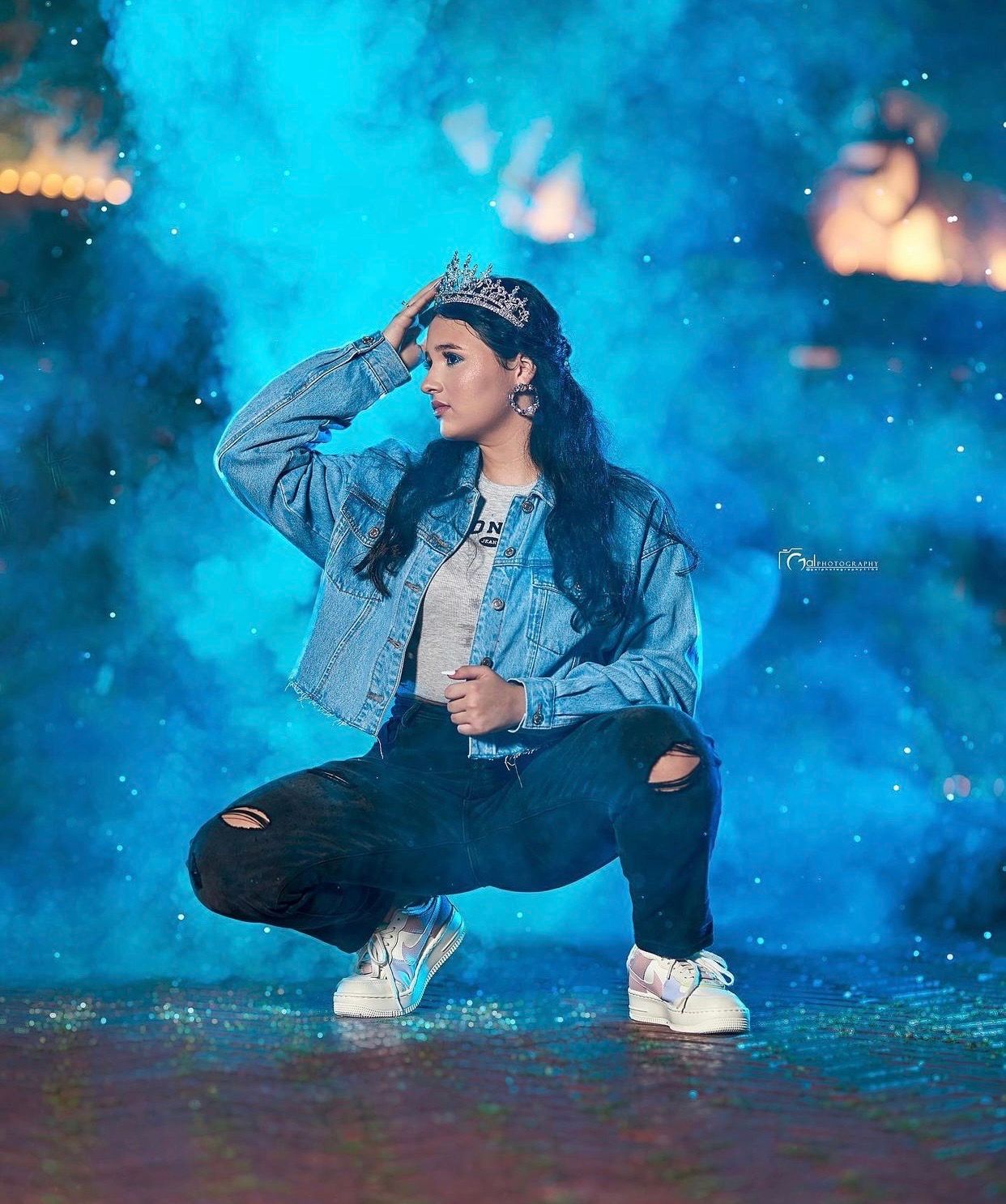 Young woman in denim jacket and crown poses dramatically against blue smoke background.