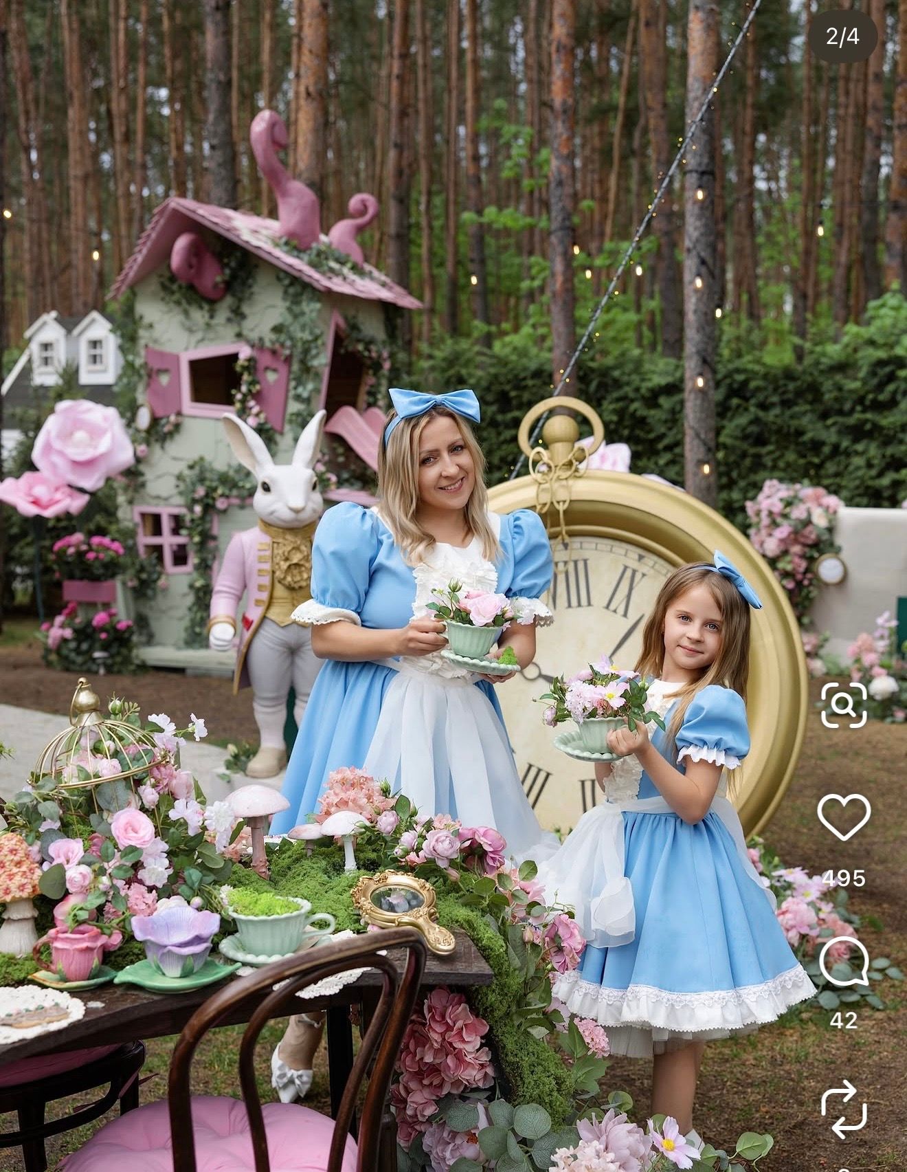Woman and girl in blue Alice in Wonderland dresses at a whimsical forest tea party with a giant clock, white rabbit statue and pink flowers.