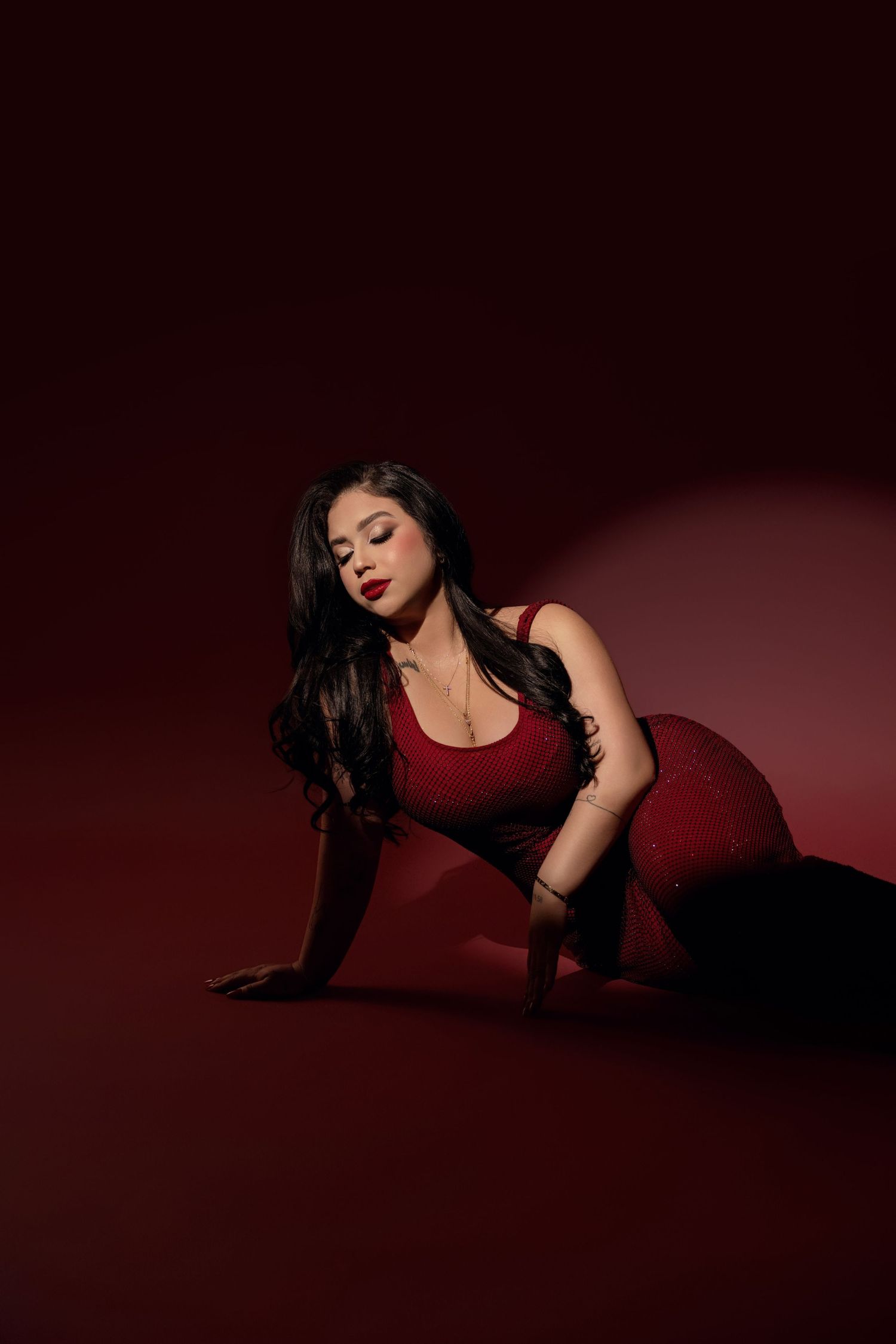 Woman with long dark hair and red lipstick poses on the floor in a red top and pants against a moody dark red studio backdrop.