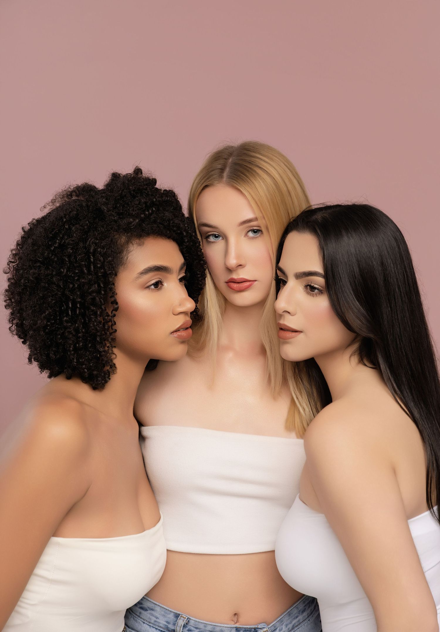 Three women with curly black, straight blonde, and long dark hair pose closely together in white tube tops against a dusty pink background.