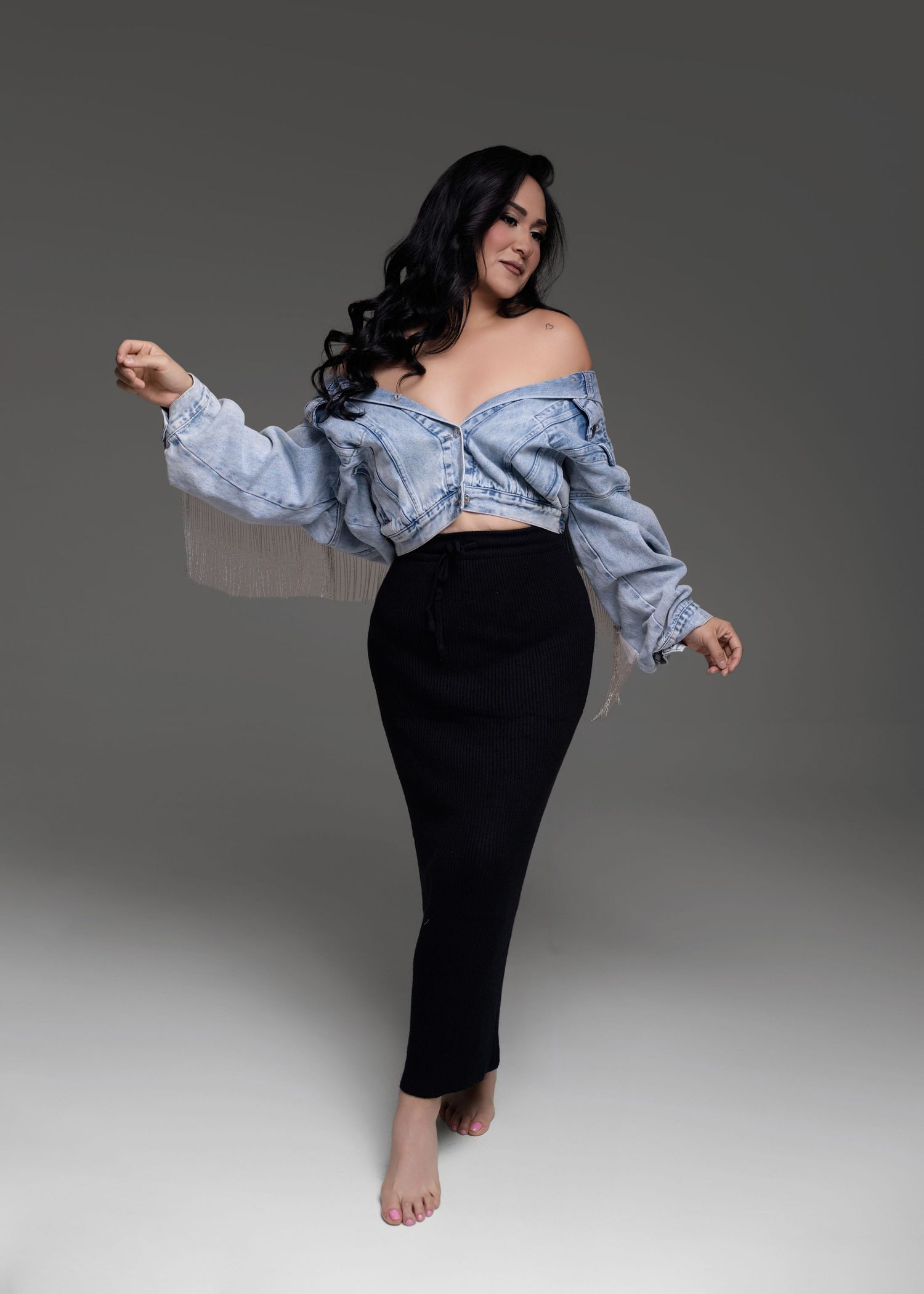 Woman with long wavy dark hair poses barefoot in an off-shoulder fringed denim jacket and black maxi skirt against a gray studio backdrop.