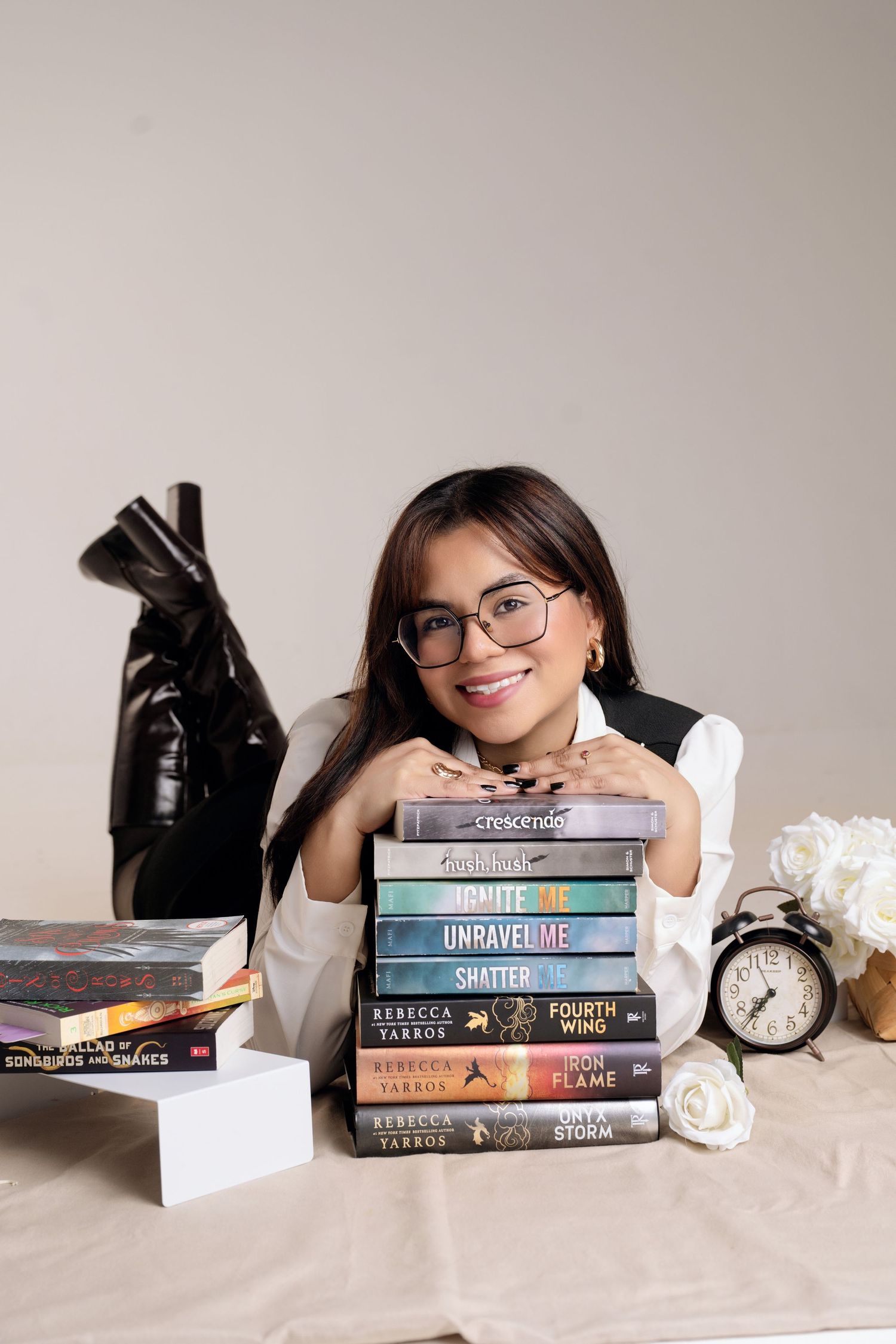 Smiling woman with glasses rests her chin on a stack of Rebecca Yarros books beside a vintage alarm clock and white flowers.