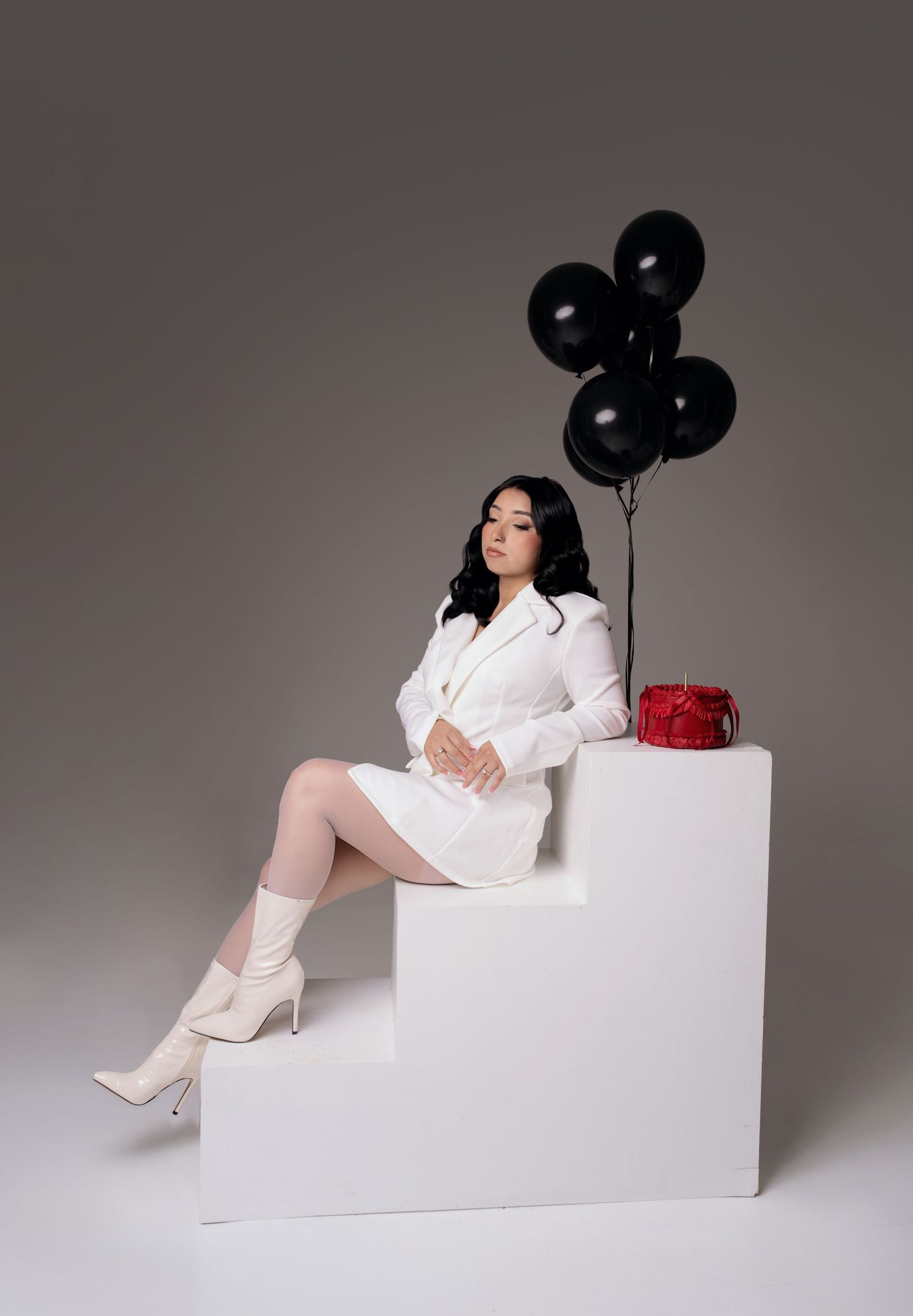 Woman in white blazer dress and boots sits on white steps with black balloons and red gift box.
