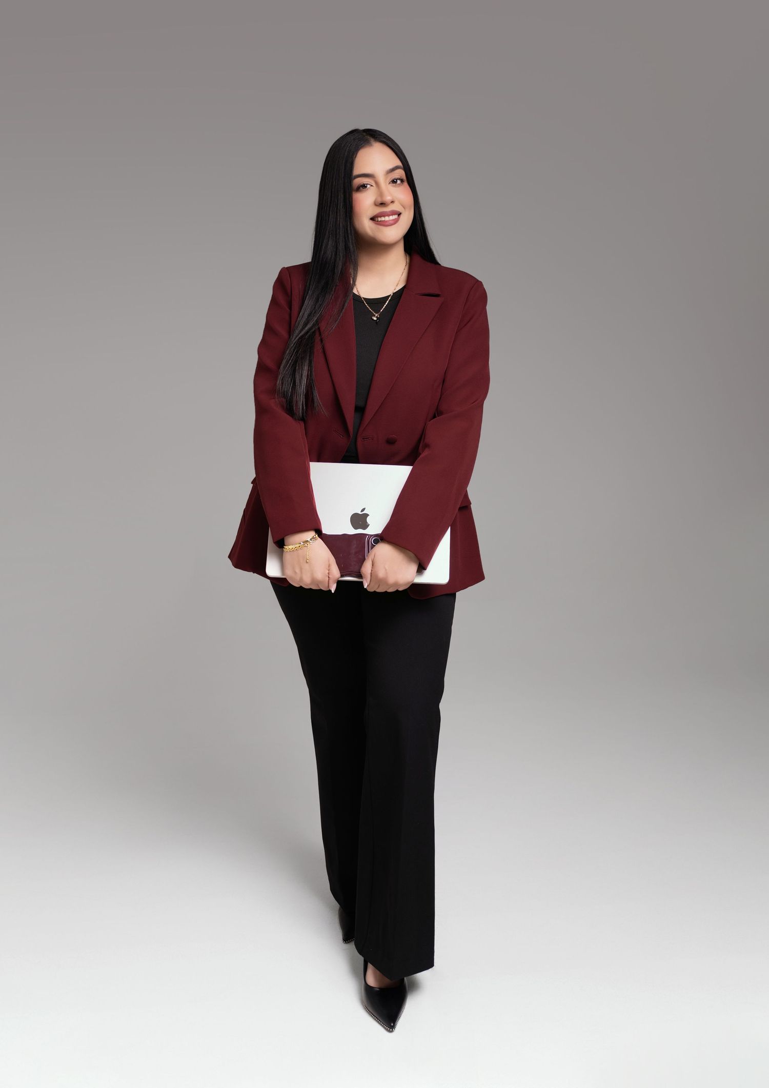 Professional woman in burgundy blazer and black pants holding Apple laptop, posing confidently in studio setting.