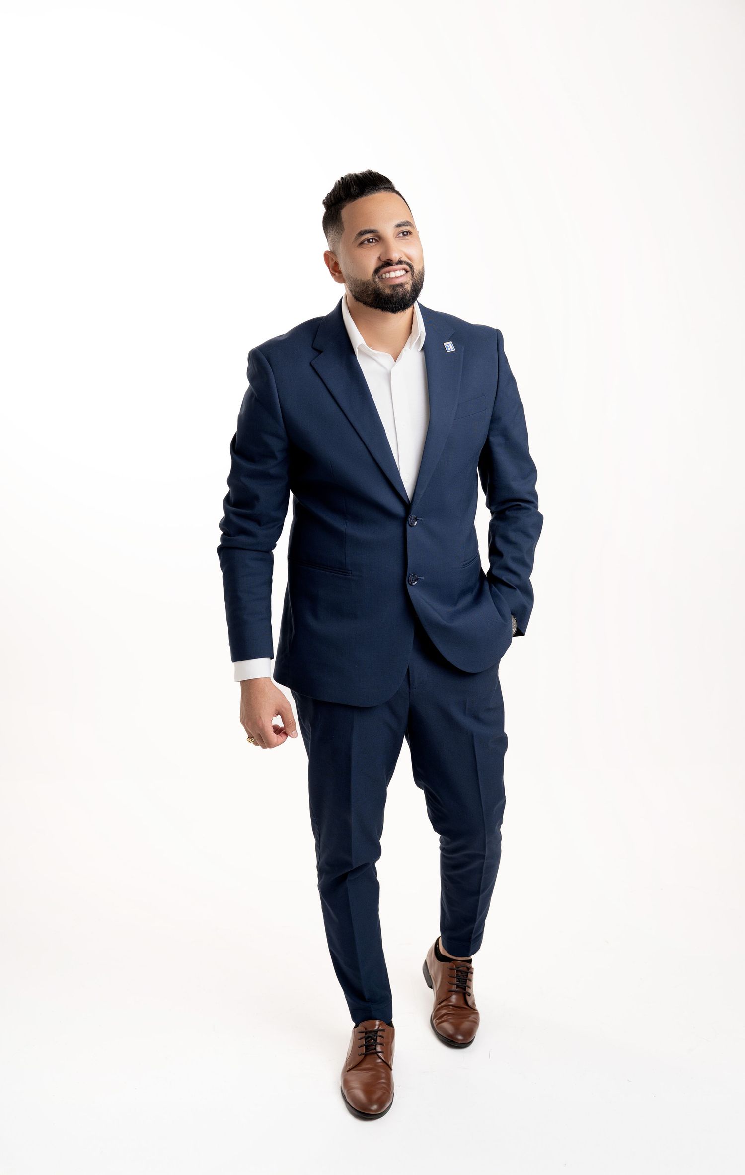 Man in navy blue suit and brown shoes walking confidently against white background.