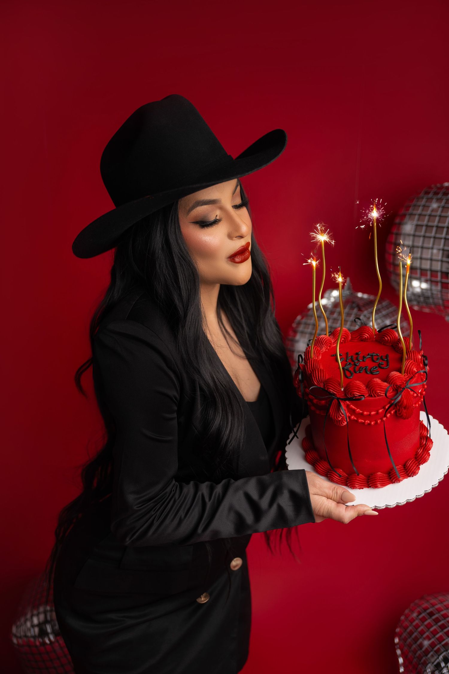 Woman in black hat and dress holding a red birthday cake with sparklers on a red background.