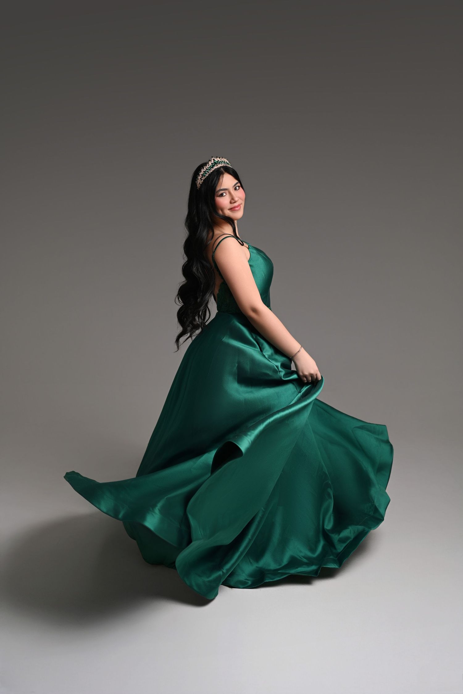 Woman in flowing emerald green satin gown and tiara poses gracefully in studio.