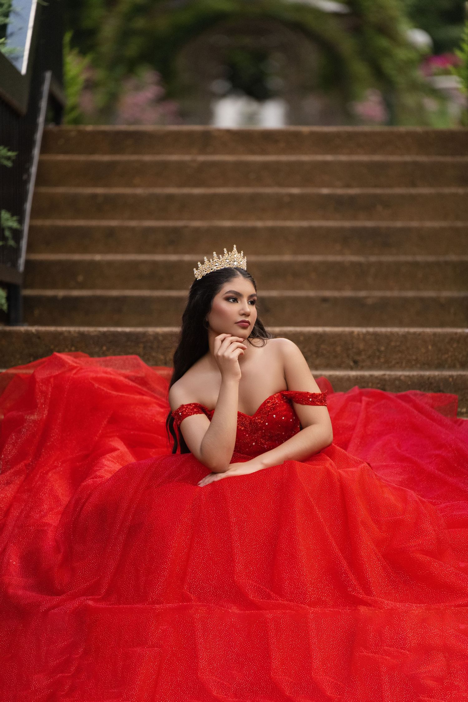 Young woman in red off-shoulder ball gown and gold crown sits gracefully on stone steps surrounded by lush greenery.