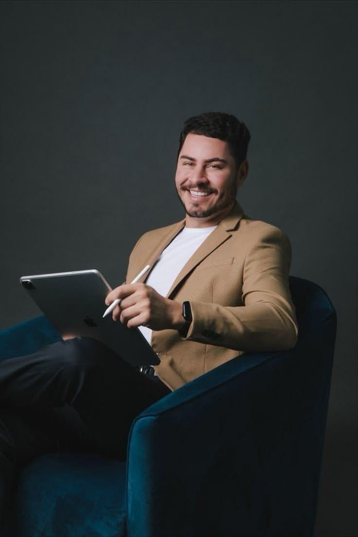 Smiling businessman in tan blazer sitting in blue velvet chair using digital tablet with stylus.