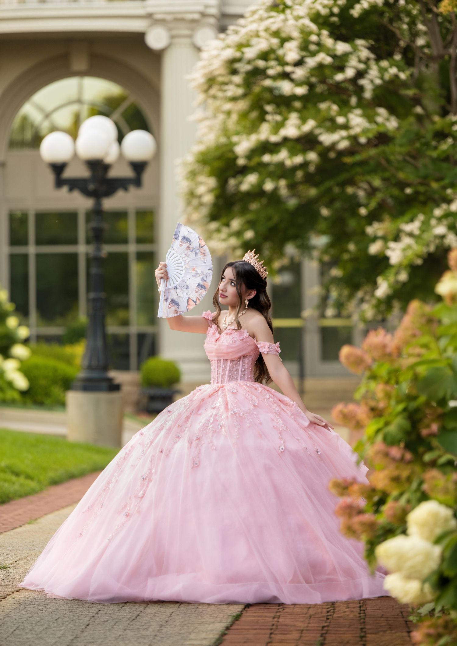 Young woman in pink ball gown and crown holds decorative fan in garden for quinceañera celebration.
