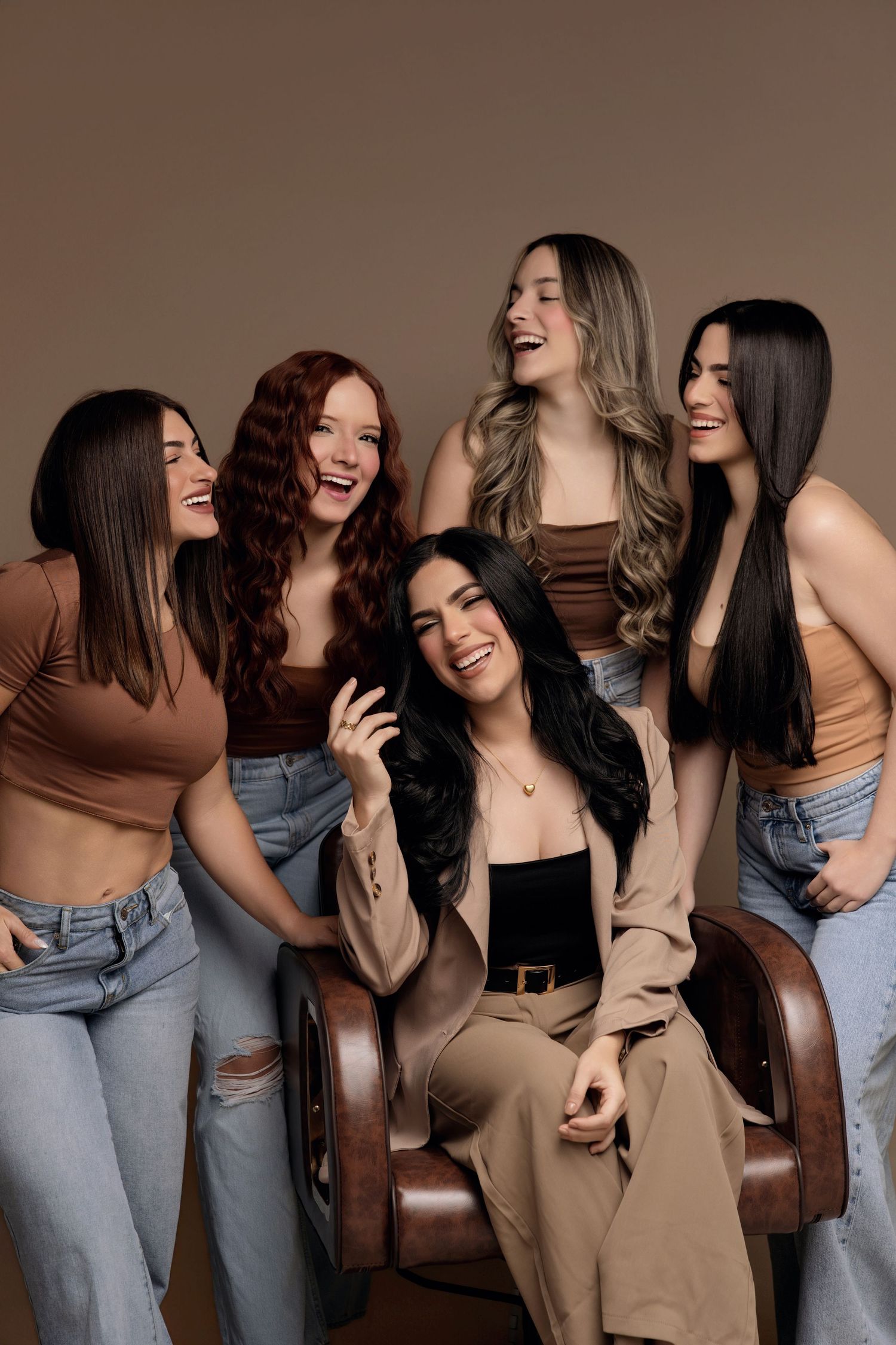 Five young women laughing and posing together in a studio, wearing neutral tones and jeans against a brown background.