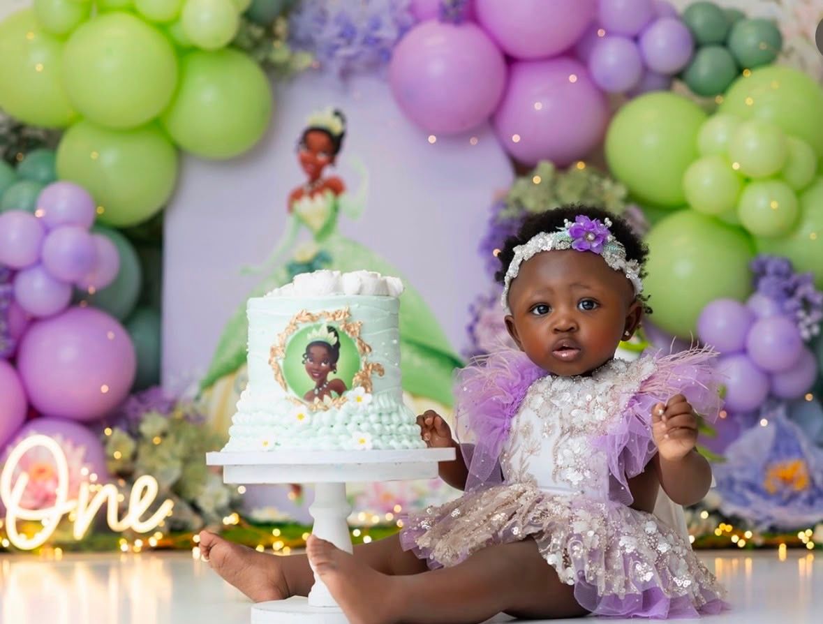 Baby girl in purple tutu dress sits with Princess Tiana first birthday cake, surrounded by green and purple balloon arch.