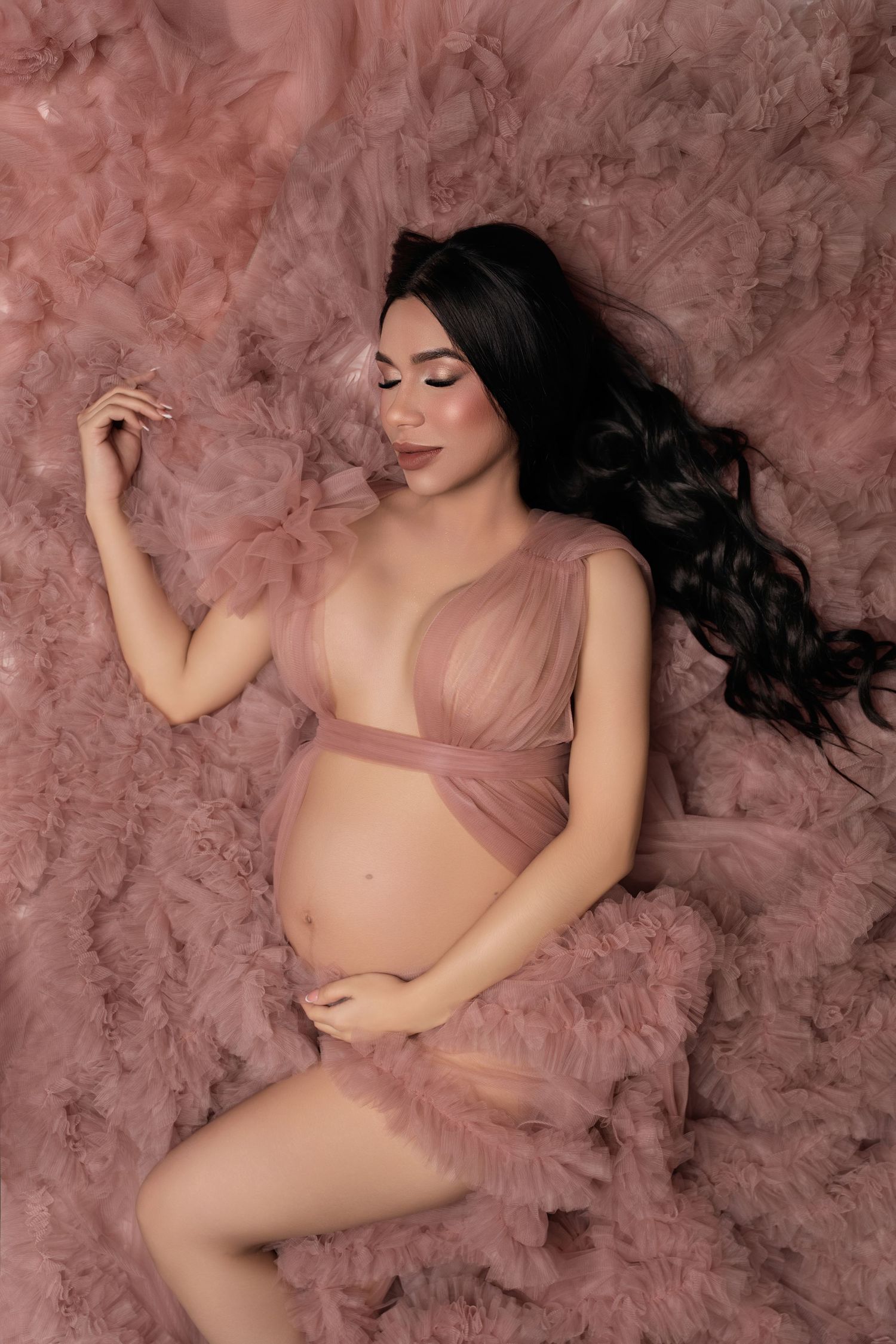 Pregnant woman in pink tulle robe lying gracefully on fluffy pink fabric for a maternity photoshoot.