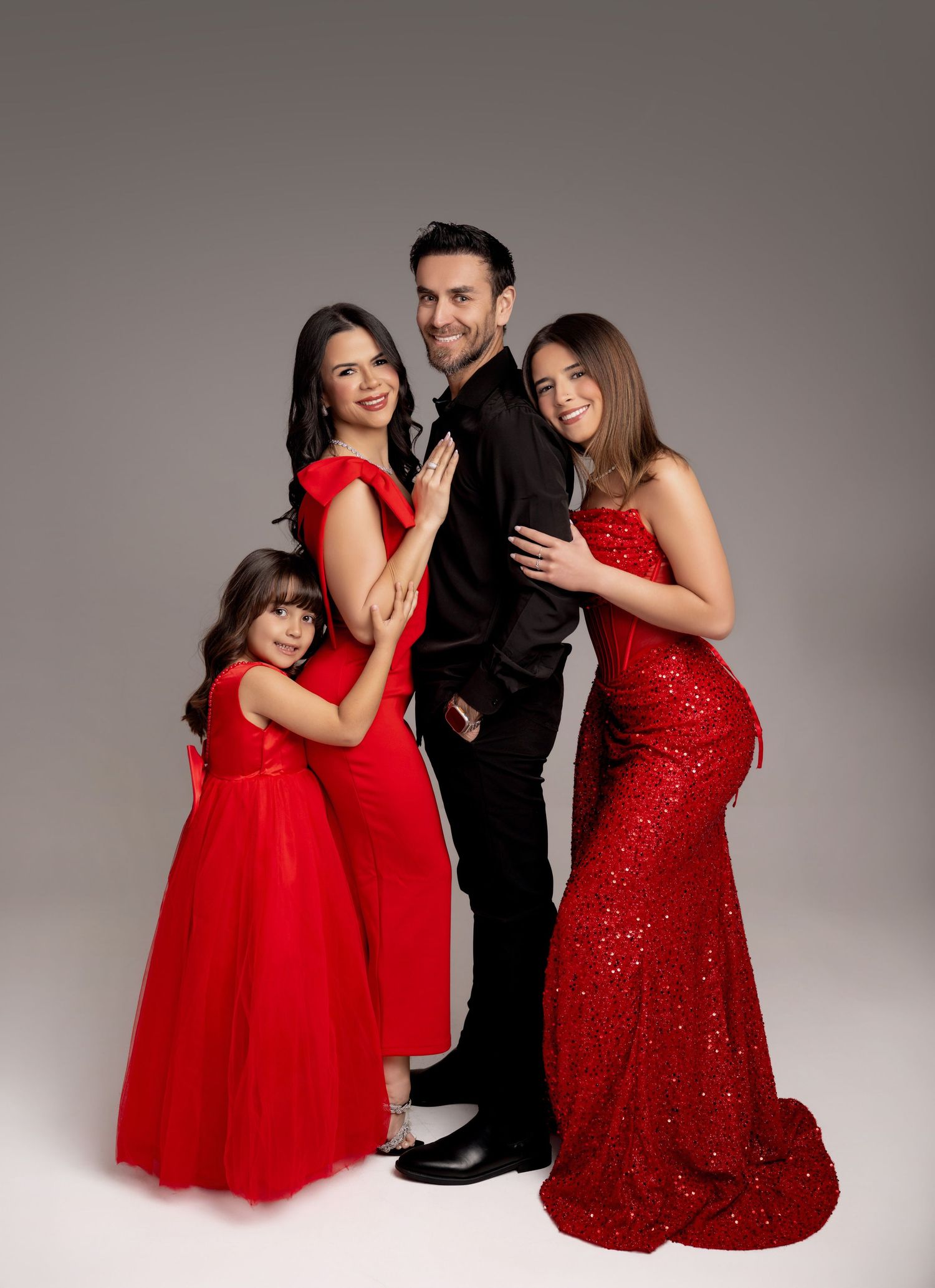 Family of four posing in studio, three in red dresses and one in black suit, elegant holiday portrait.