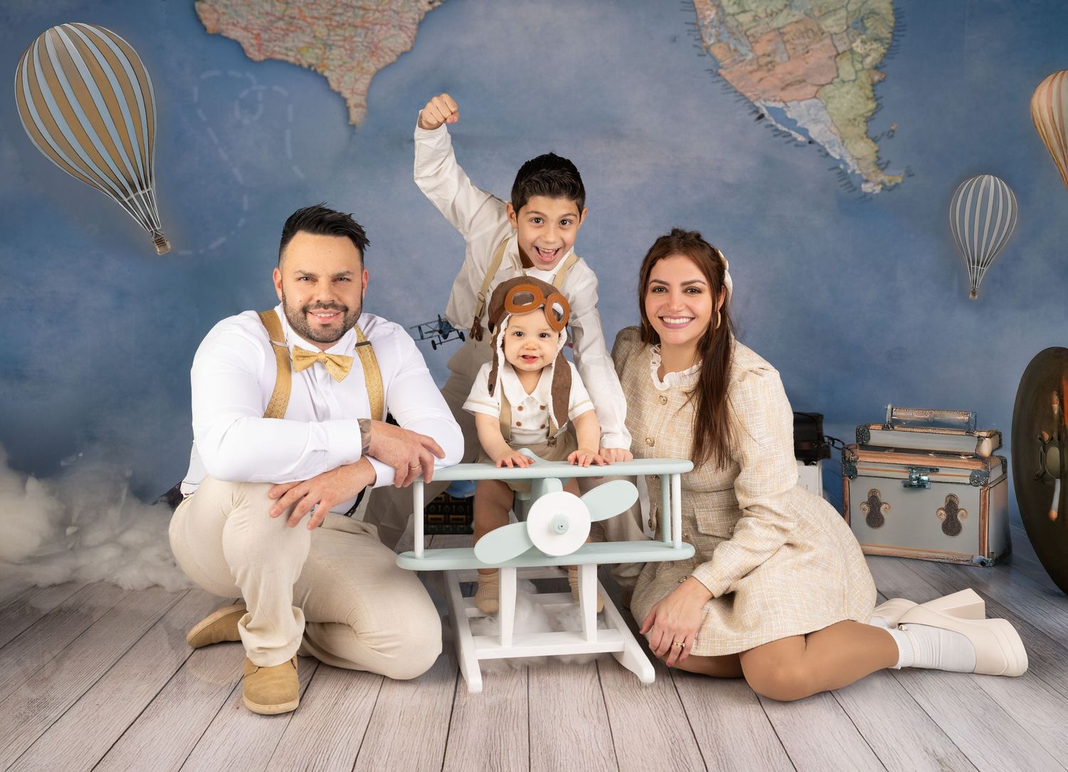 Family of four posing in front of a world map backdrop with hot air balloons, featuring a toy airplane and vintage suitcases.