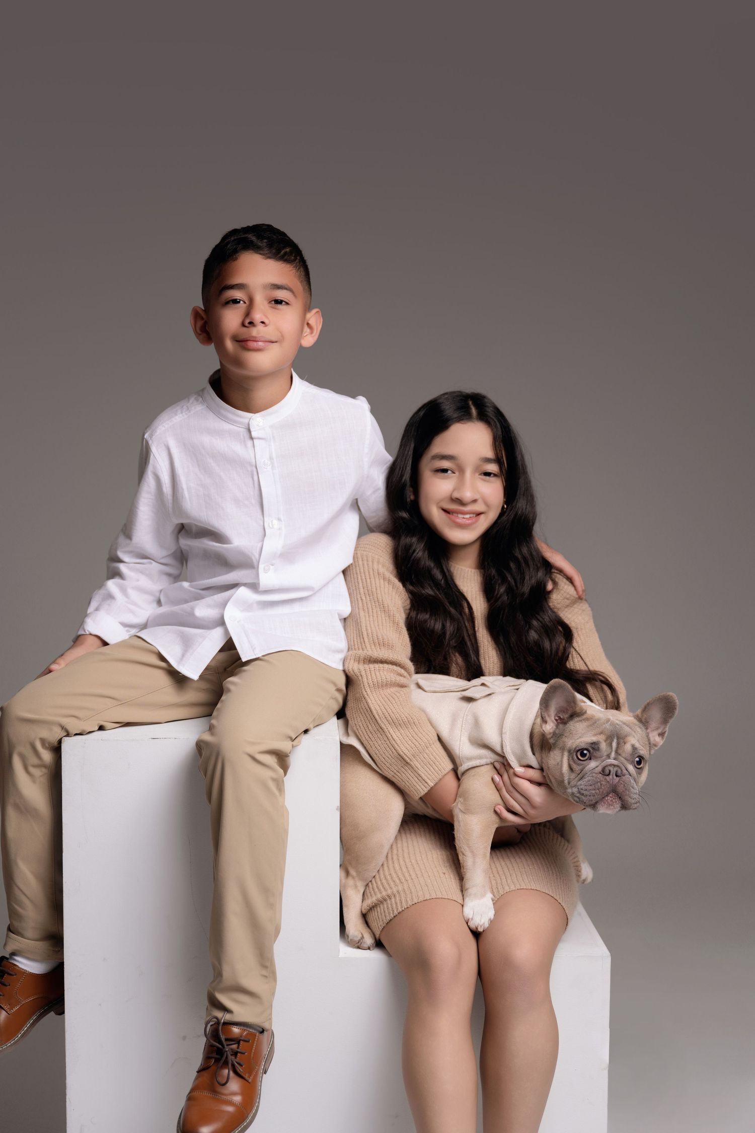 Two children and a French Bulldog pose on a white cube against a gray backdrop in stylish neutral outfits.