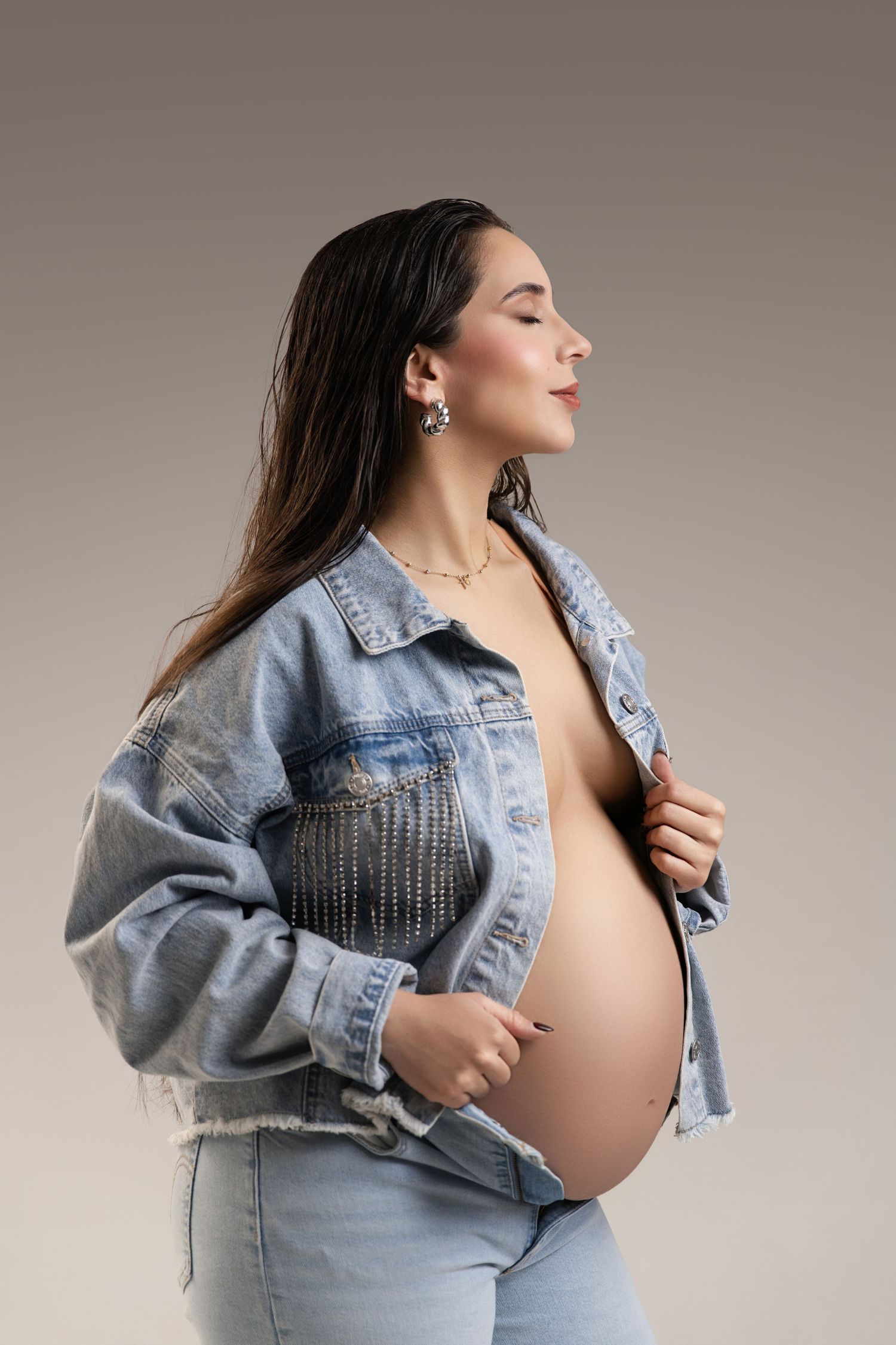 Pregnant woman in denim jacket and jeans posing sideways, showing baby bump, with long dark hair and elegant earrings.