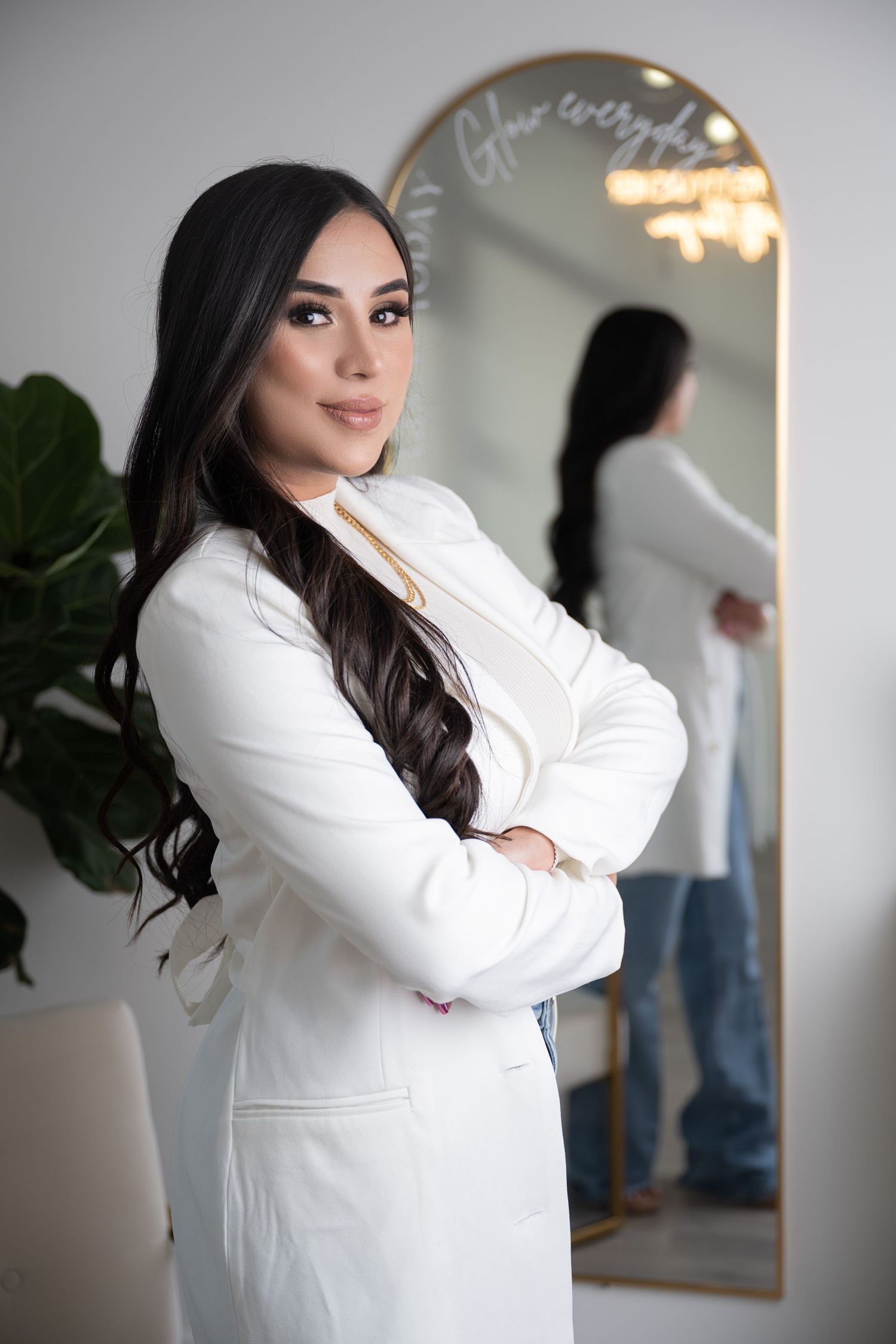Professional in white blazer poses confidently near decorative mirror and plant.