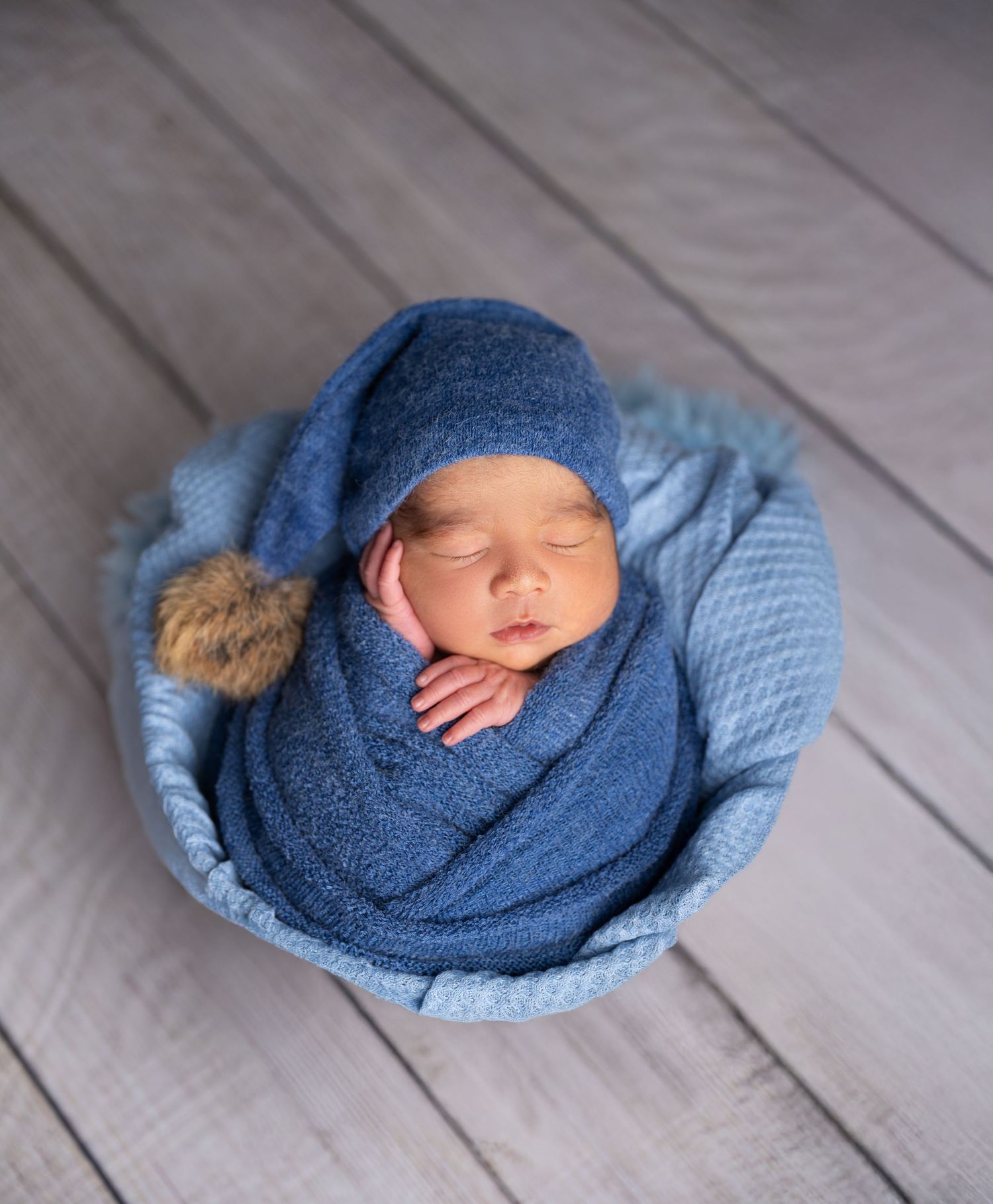 Cozy newborn portrait with blue knit wrap and pom-pom hat on wooden backdrop.