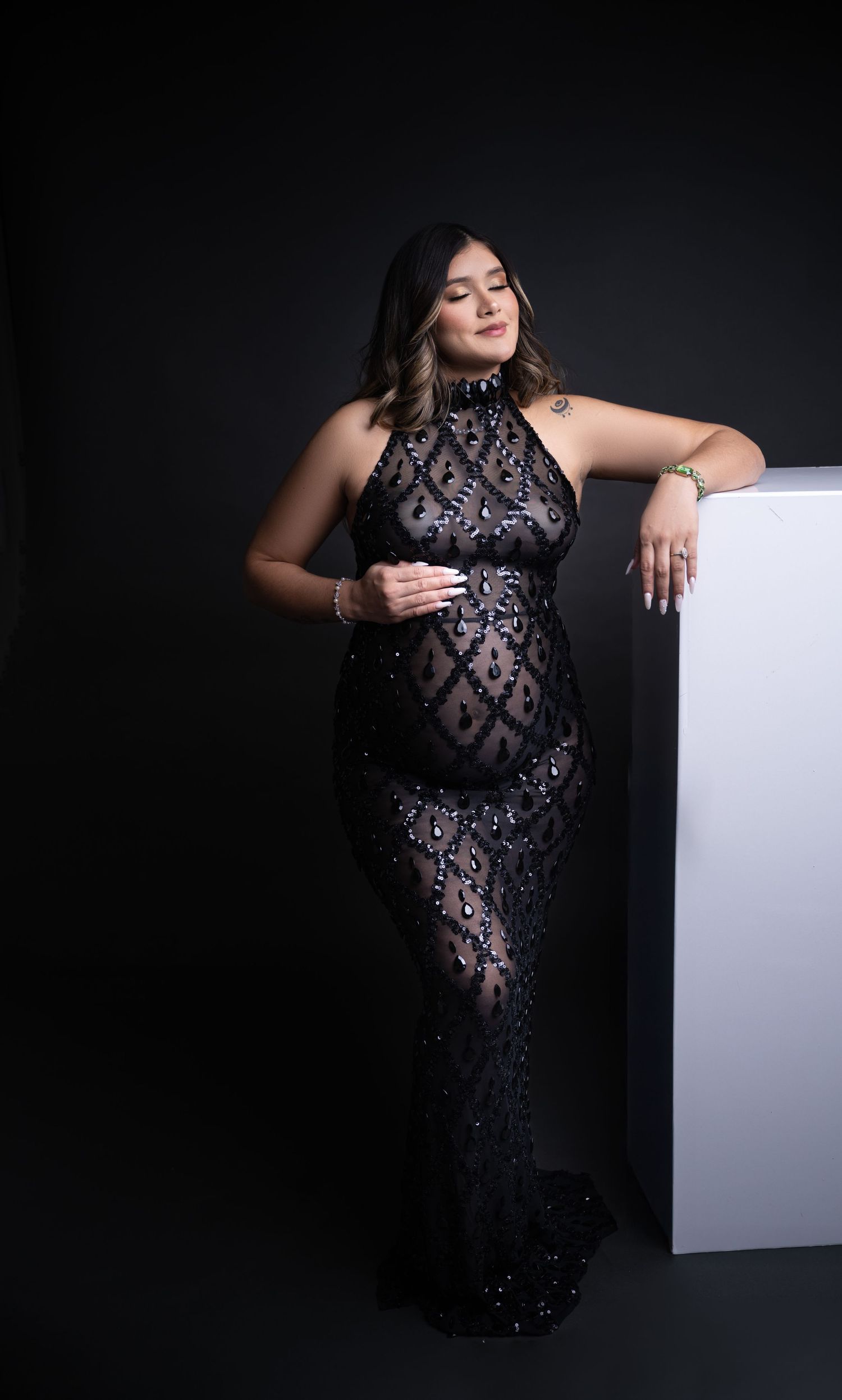 Woman in sequined black gown leans gracefully against white cube in studio setting.