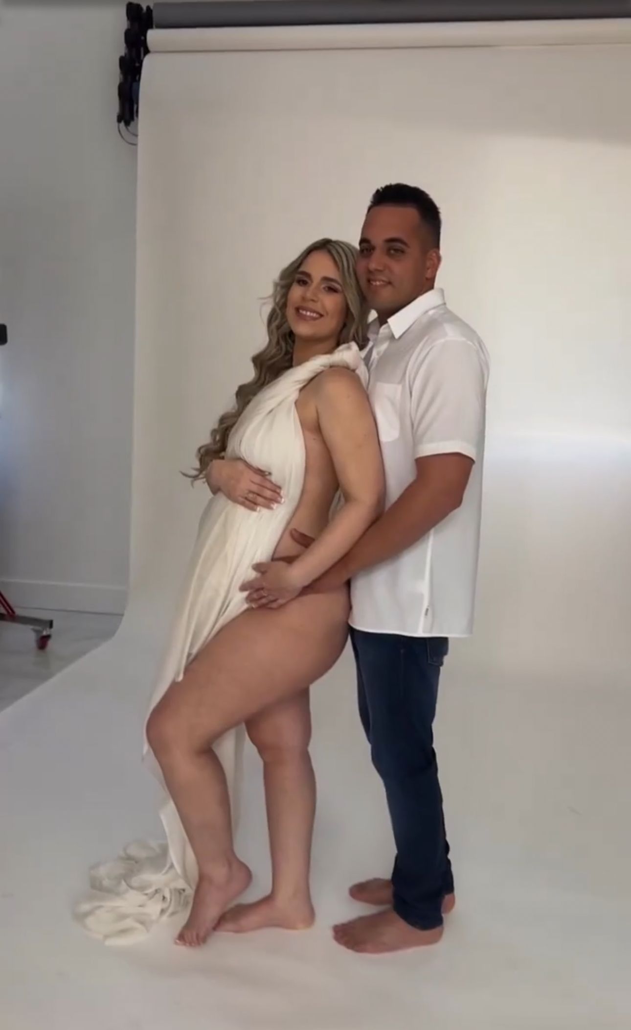 Pregnant woman in white draped fabric poses with partner in white shirt and jeans during maternity photoshoot.
