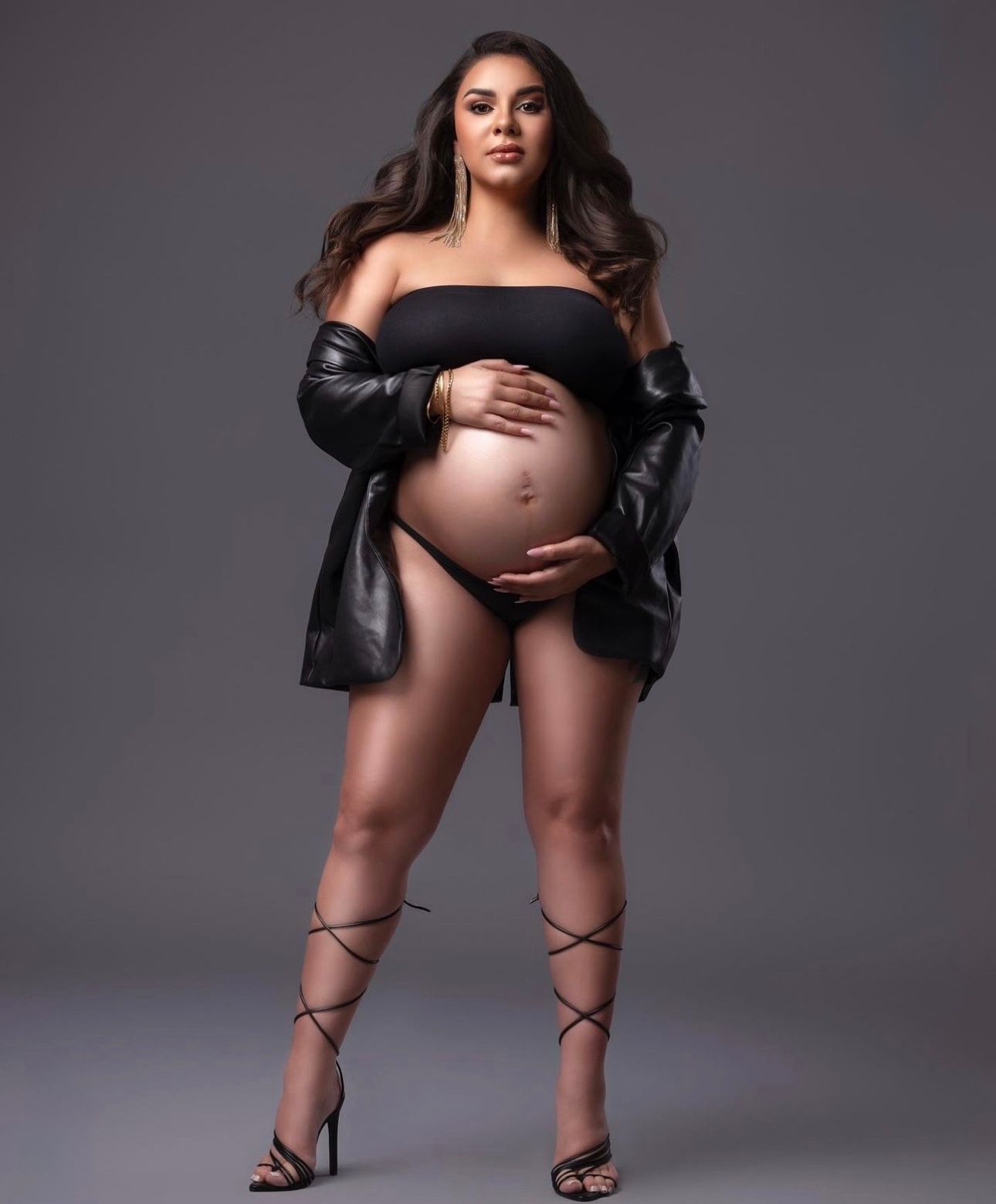 Pregnant woman in black strapless bodysuit, leather jacket, and strappy heels posing for a maternity photoshoot.