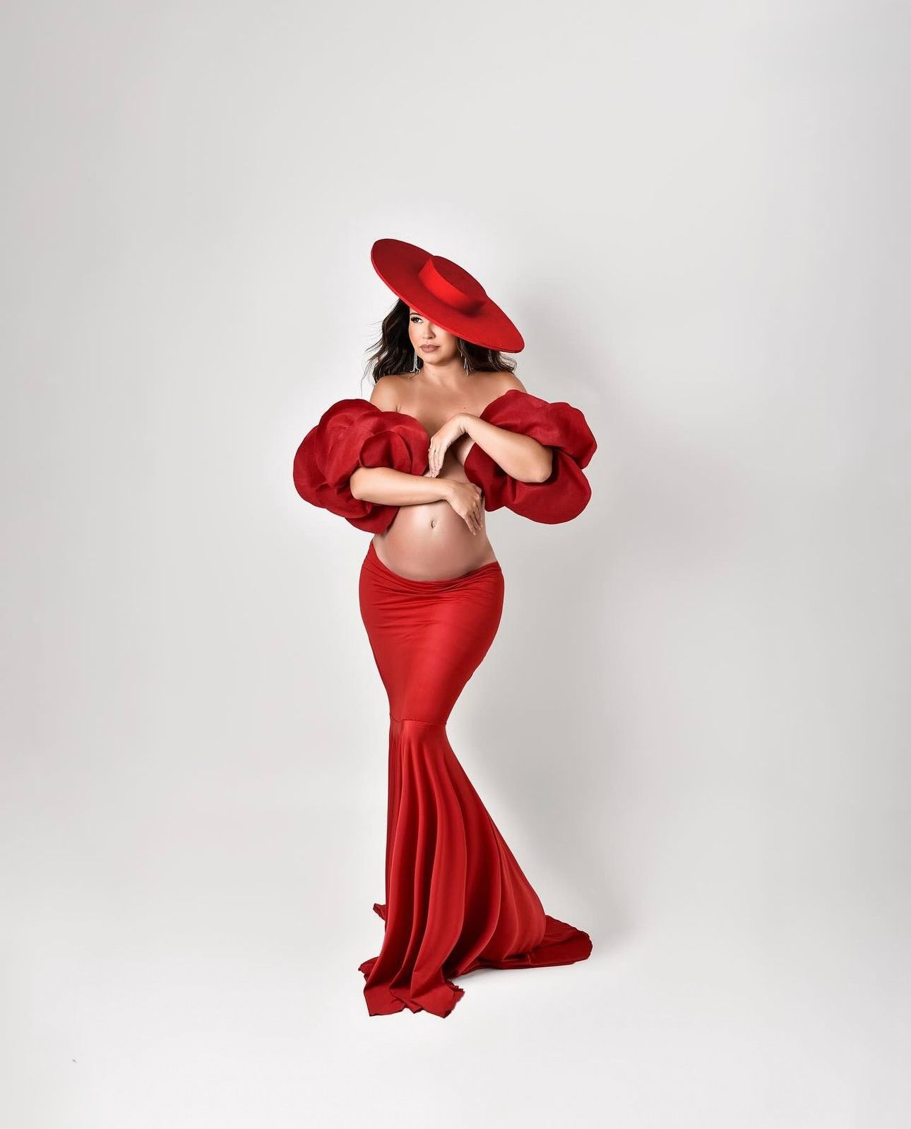 Pregnant woman in red off-shoulder puff sleeve crop top, matching mermaid skirt and beret hat posing against white background.