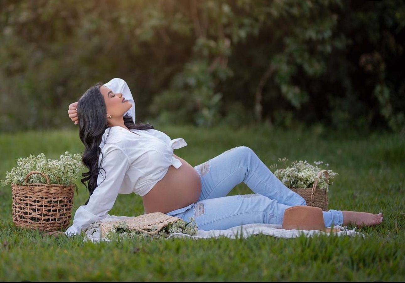 Pregnant woman in white shirt and jeans relaxing on grass during outdoor maternity photoshoot with flower baskets.