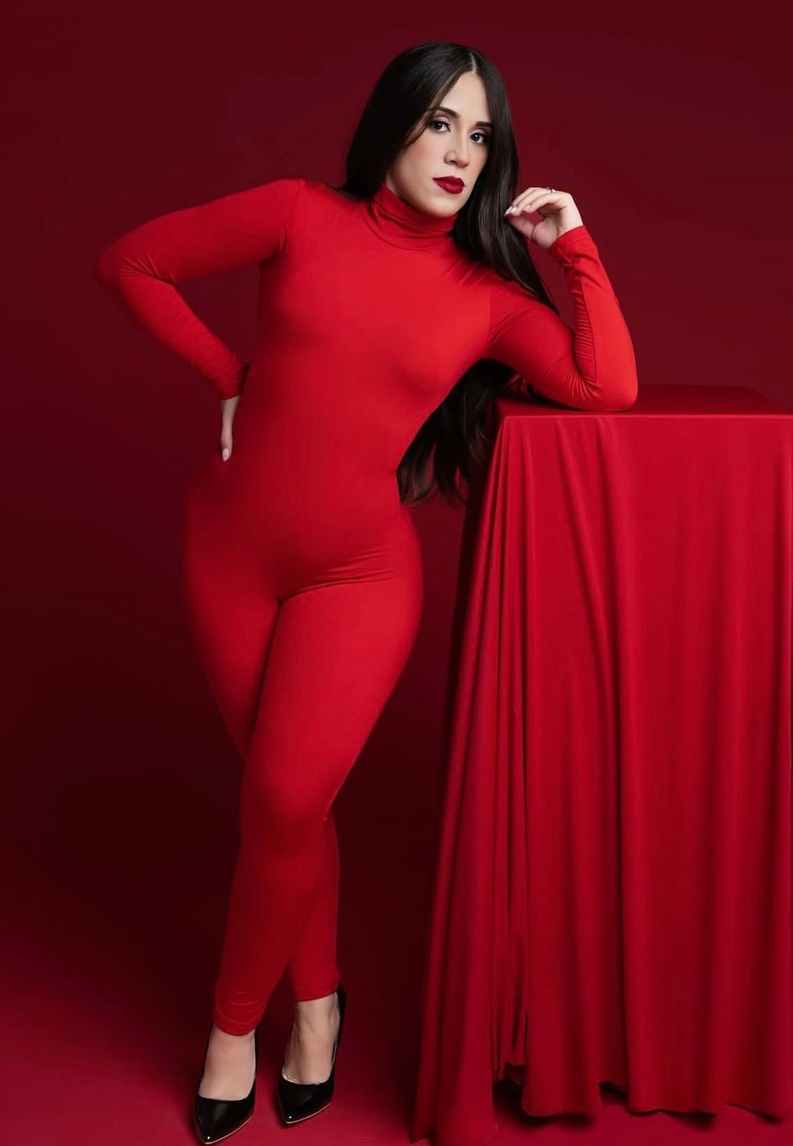 Woman in red turtleneck catsuit poses confidently against red background, wearing black heels.