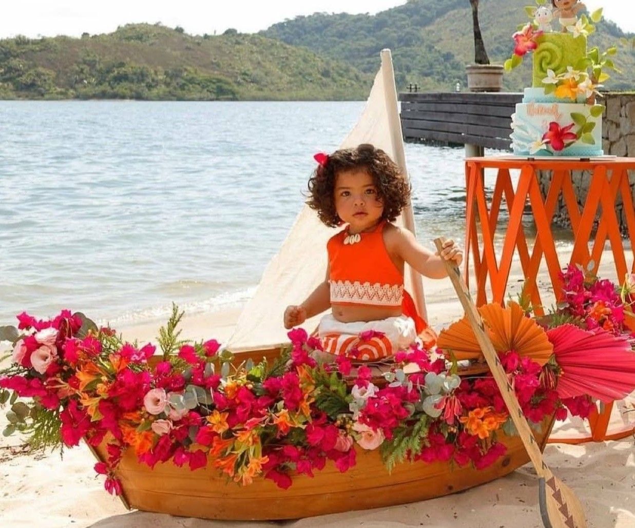 Young girl dressed as Moana sits in a flower-decorated canoe on a tropical beach with lush greenery.