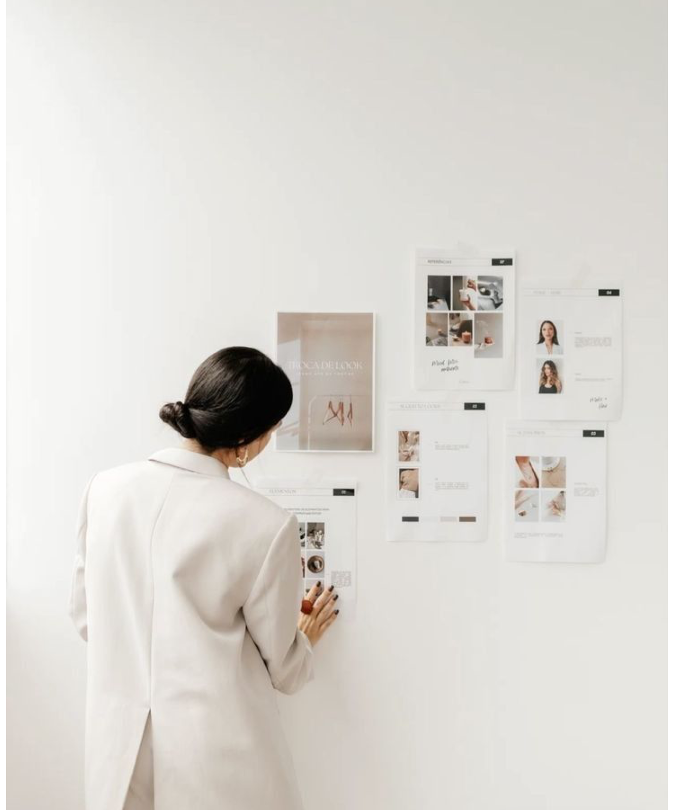 Woman in white blazer arranging design mood board with photos and layouts on a white wall.